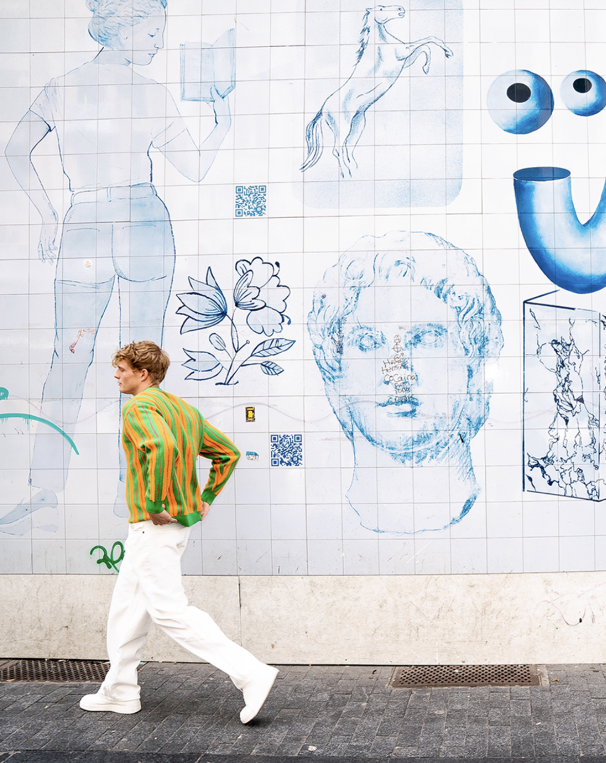A man wearing a colorful striped jacket and white pants walking past a large tiled mural featuring line sketches of a woman reading a book, a horse, a face, a butterfly, eyeballs, and QR codes.