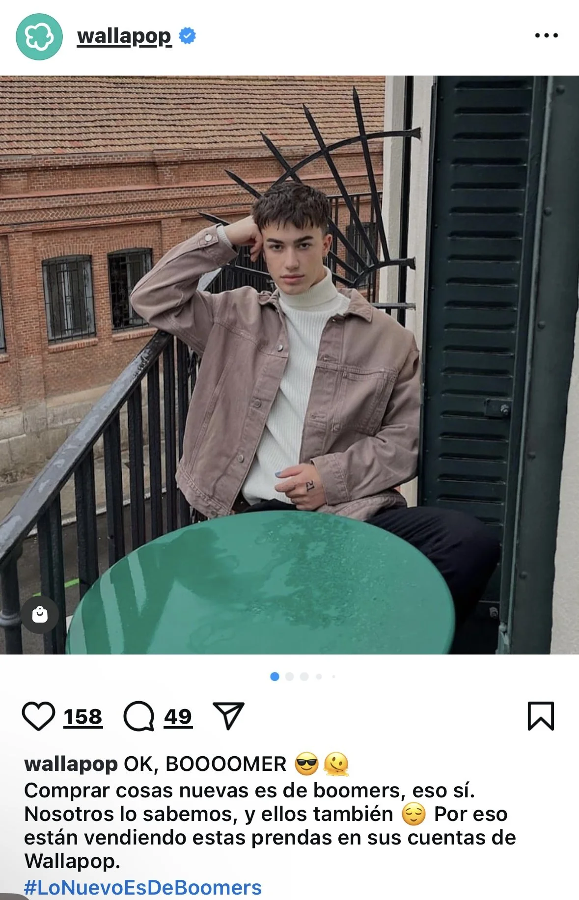 A young man with dark hair sitting on a balcony railing, leaning against a wall with black shutters, wearing a beige jacket and a white turtleneck sweater, with a green round table in front of him, and a brick building in the background.