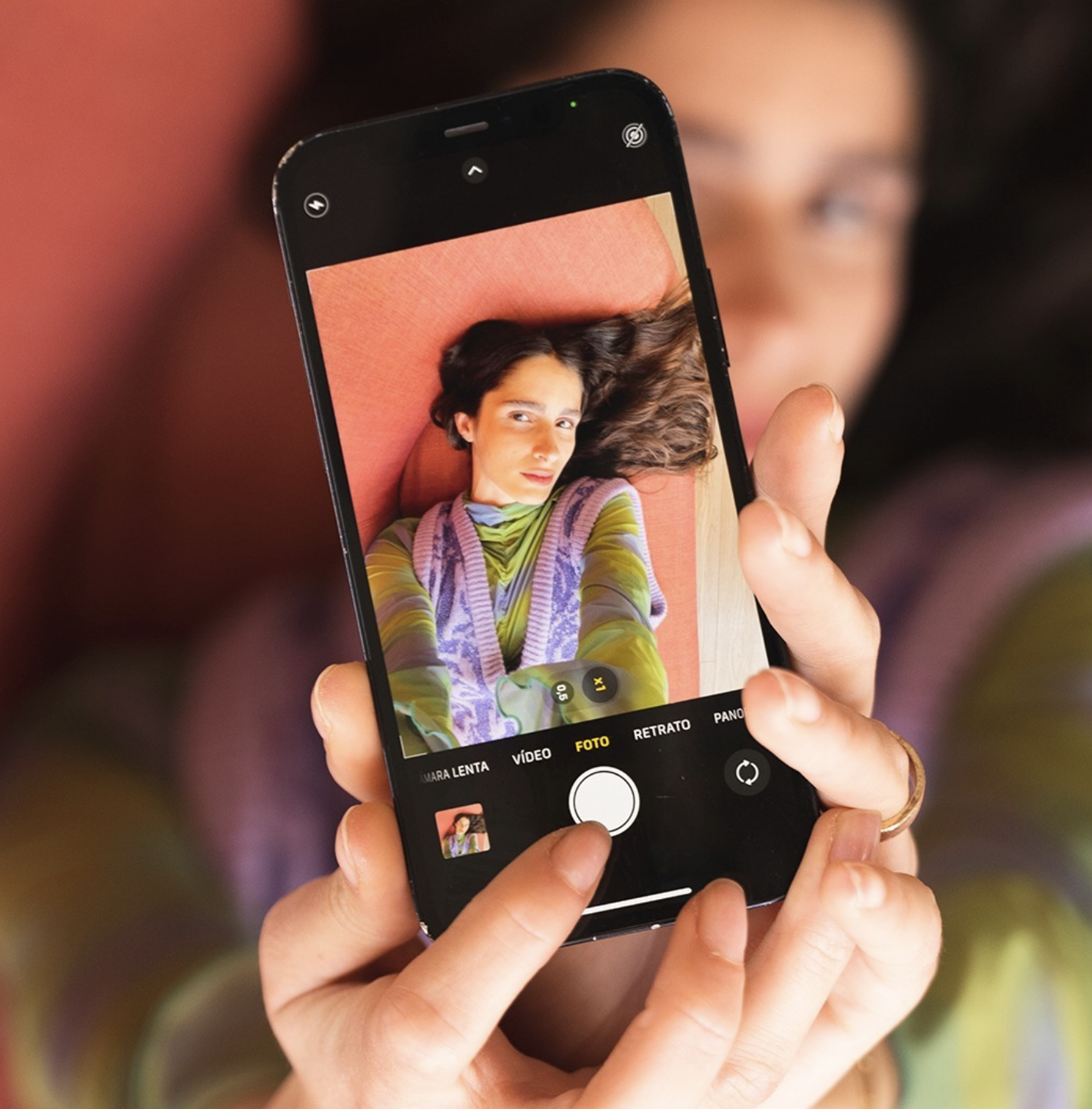 Person taking a selfie with a smartphone, showing a woman with dark hair lying on a pinkish-orange surface, wearing a colorful striped sweater.