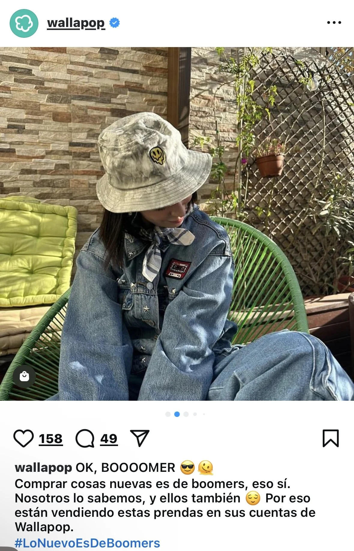 A woman sitting outdoors on a green circular chair, wearing a tie-dye bucket hat, a denim jacket, and jeans, with a black and white checkered scarf around her neck, against a backdrop of a stone wall, a green hammock, and potted plants.