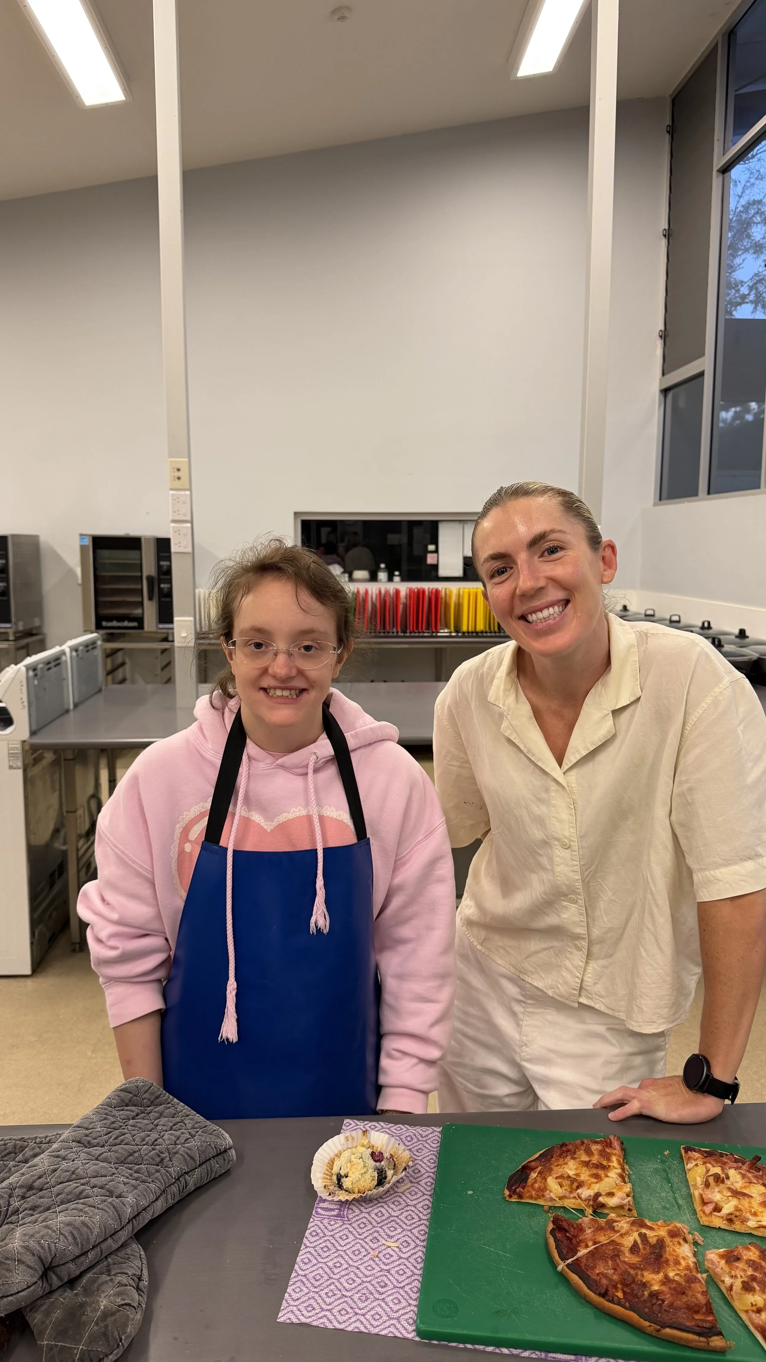 A smiling woman and girl stand in a kitchen near a table with sliced pizza and a cupcake, with kitchen appliances and colorful items in the background.