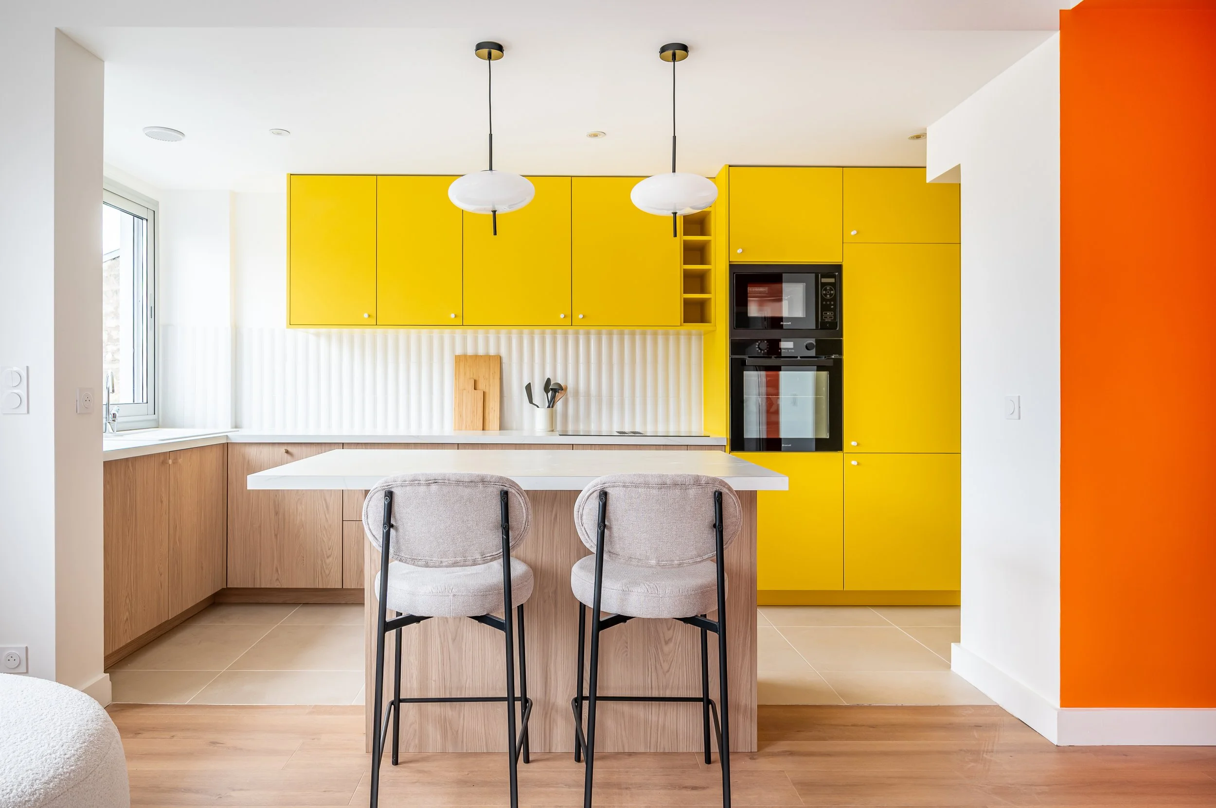 Une cuisine moderne avec des armoires jaunes, un mur blanc avec un revêtement en lamelles verticales, une table blanche, deux chaises avec coussin gris, une fenêtre, et un mur orange à droite.