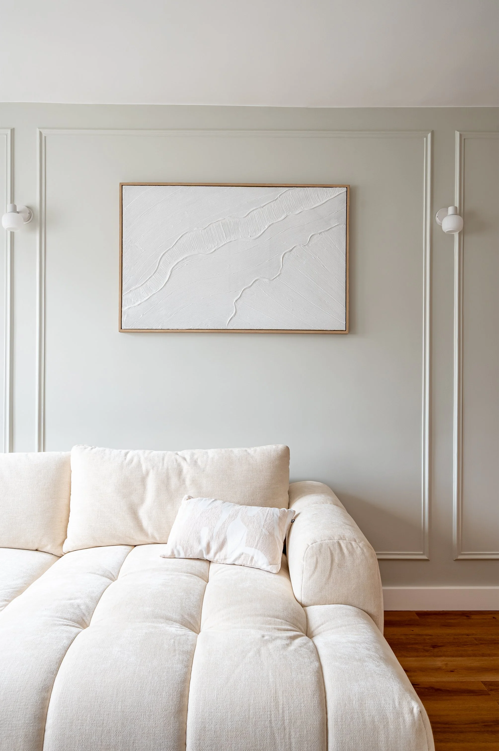 Un canapé beige avec des coussins blancs dans un salon épuré, au mur une œuvre d'art abstracte encadrée. La pièce est décorée de moulures blanches, avec des lampes murales blanches, et un sol en bois.