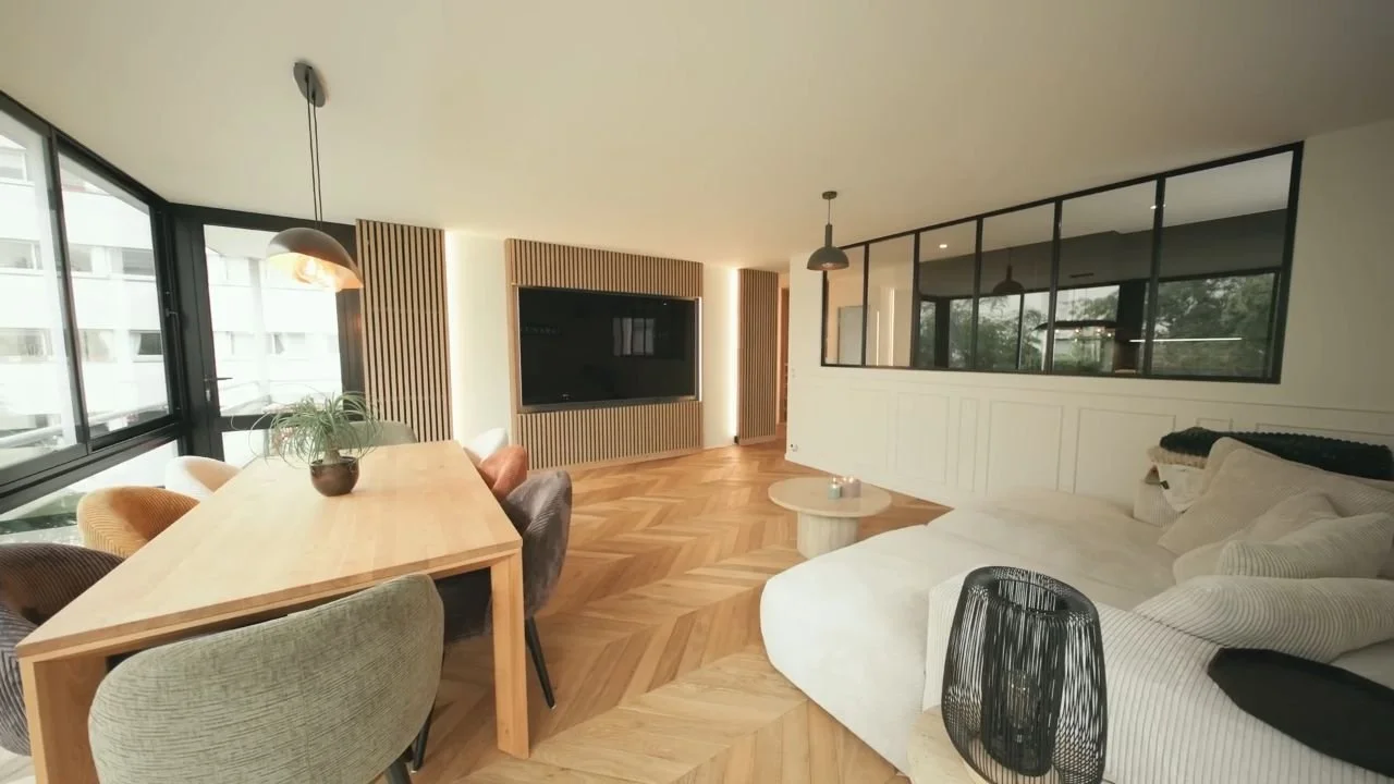 Salon moderne avec canapé blanc, table basse ronde, télévision murale encadrée de bois, et table à manger en bois avec plusieurs chaises. Grandes fenêtres avec vue sur l'extérieur, sol en parquet en chevrons.