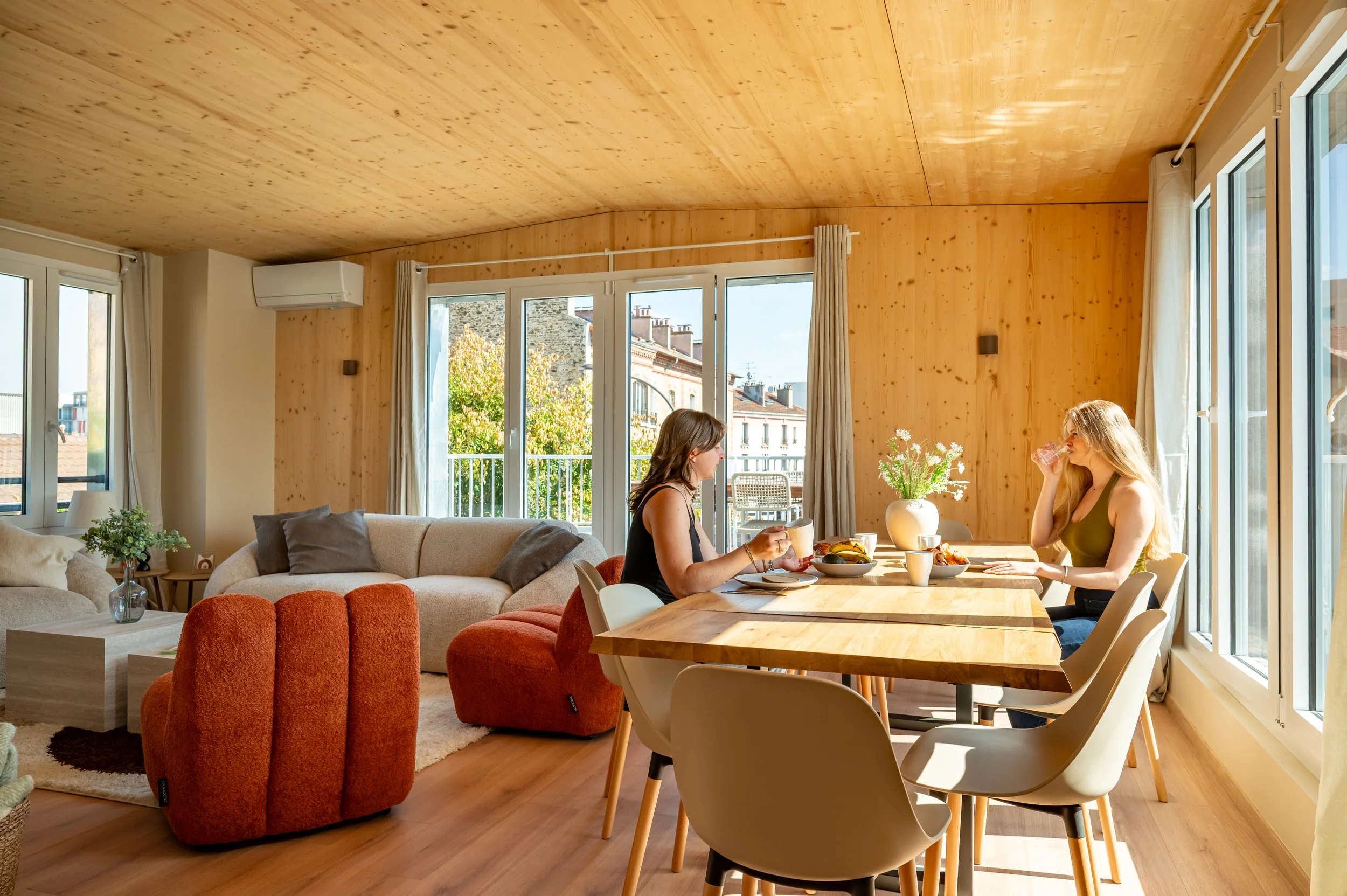 Deux femmes mangent un petit déjeuner dans une salle lumineuse avec de grandes fenêtres et un plafond en bois.