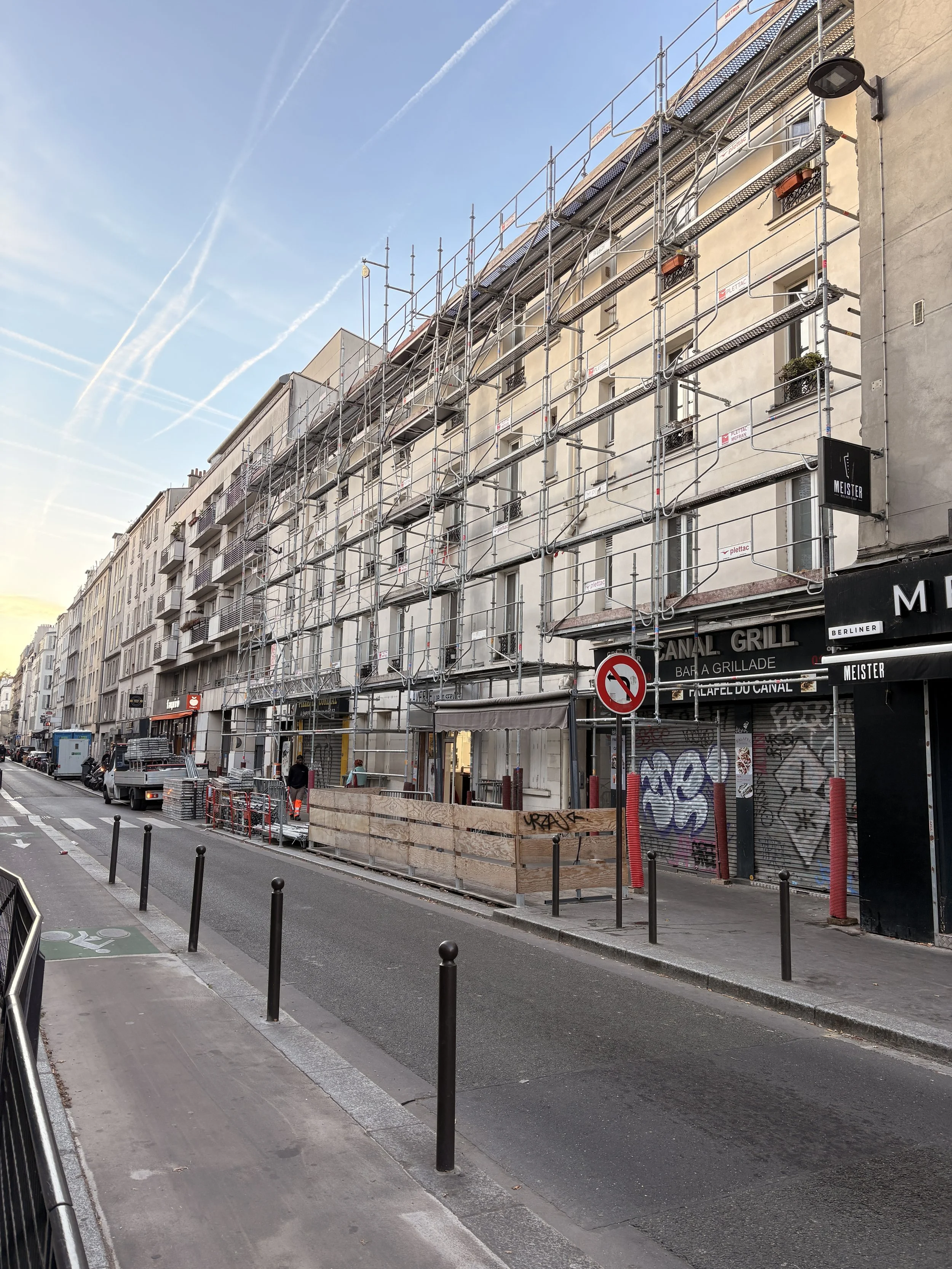 Une rue urbaine avec des bâtiments en construction, des échafaudages et des barrières de sécurité, avec un ciel bleu clair.