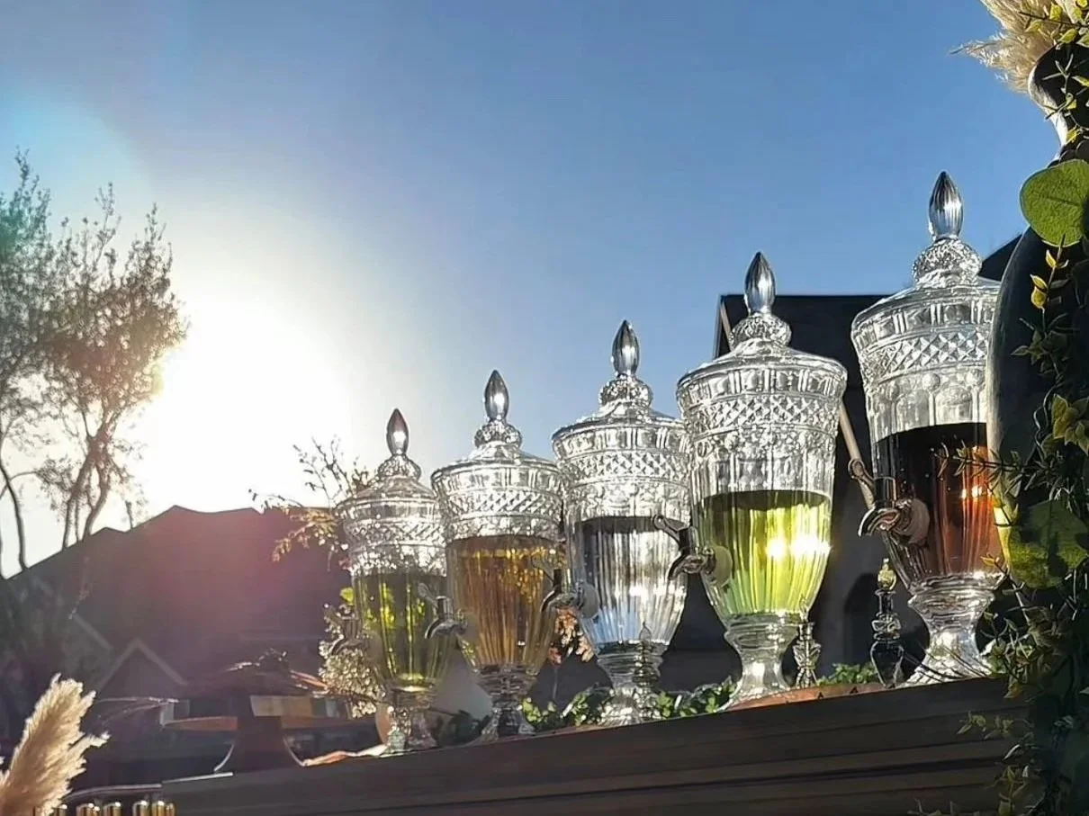 Six decorative glass jars with stoppers and spouts, filled with different colored liquids, are arranged outdoors on a ledge against a bright blue sky and sunlight.
