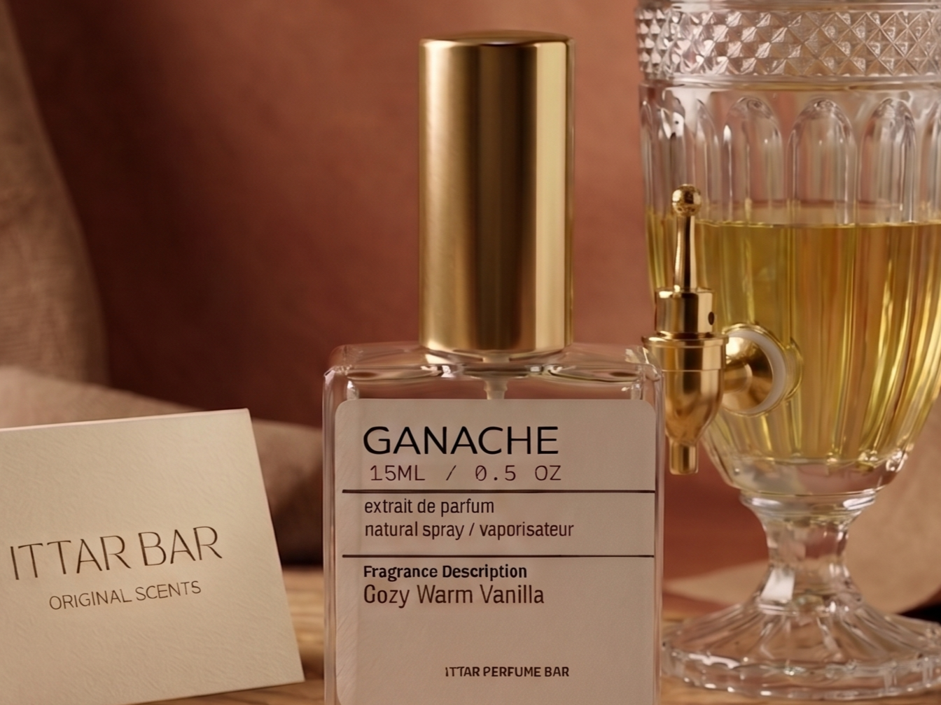 A perfume bottle labeled 'GANACHE' with a gold cap, near a glass of yellow liquid, and a card reading 'ITTAR BAR ORIGINAL SCENTS' on a wooden surface.