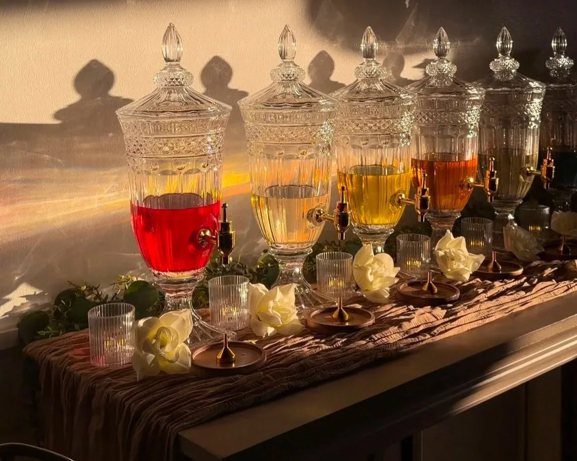 A row of crystal dispensers filled with colorful liquids, with small candles and white roses on a cloth-covered surface.