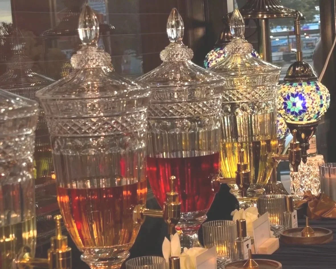 Decorative glass dispensers filled with colorful liquids on a table with small bottles and candles, with a stained glass lamp in the background.