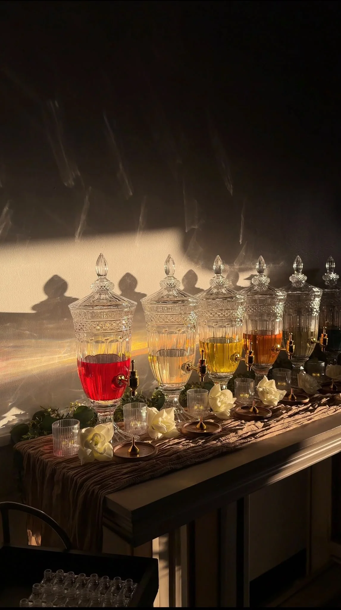 Glass dispensers with colored liquids (red, yellow, orange, white) on a table with a dark tablecloth, surrounded by white flowers, with warm ambient lighting and shadows cast on the wall behind.