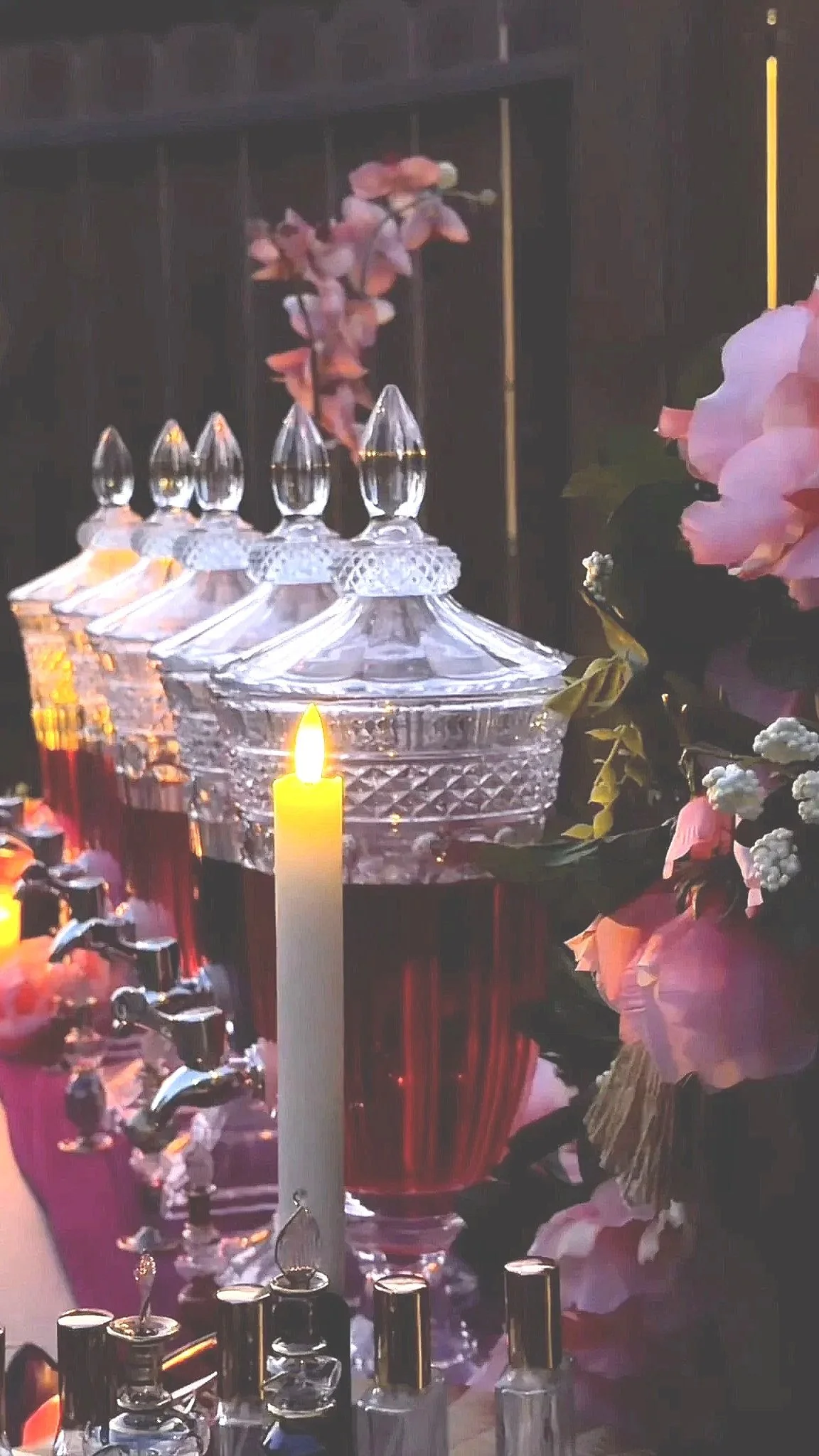 Close-up of a decorative glass container with a lid, holding a candle and surrounded by pink flowers, with perfume bottles in the foreground.