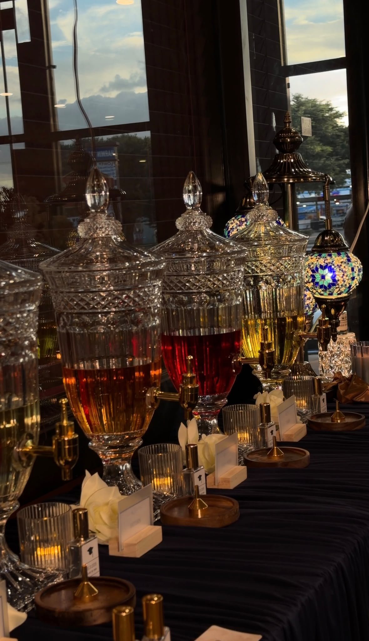 A display of ornate glass dispensers filled with colorful liquids, likely at a bar or event setup, with small tables holding candles and decorative items in front of a large window showing a cloudy sky and outdoor trees.