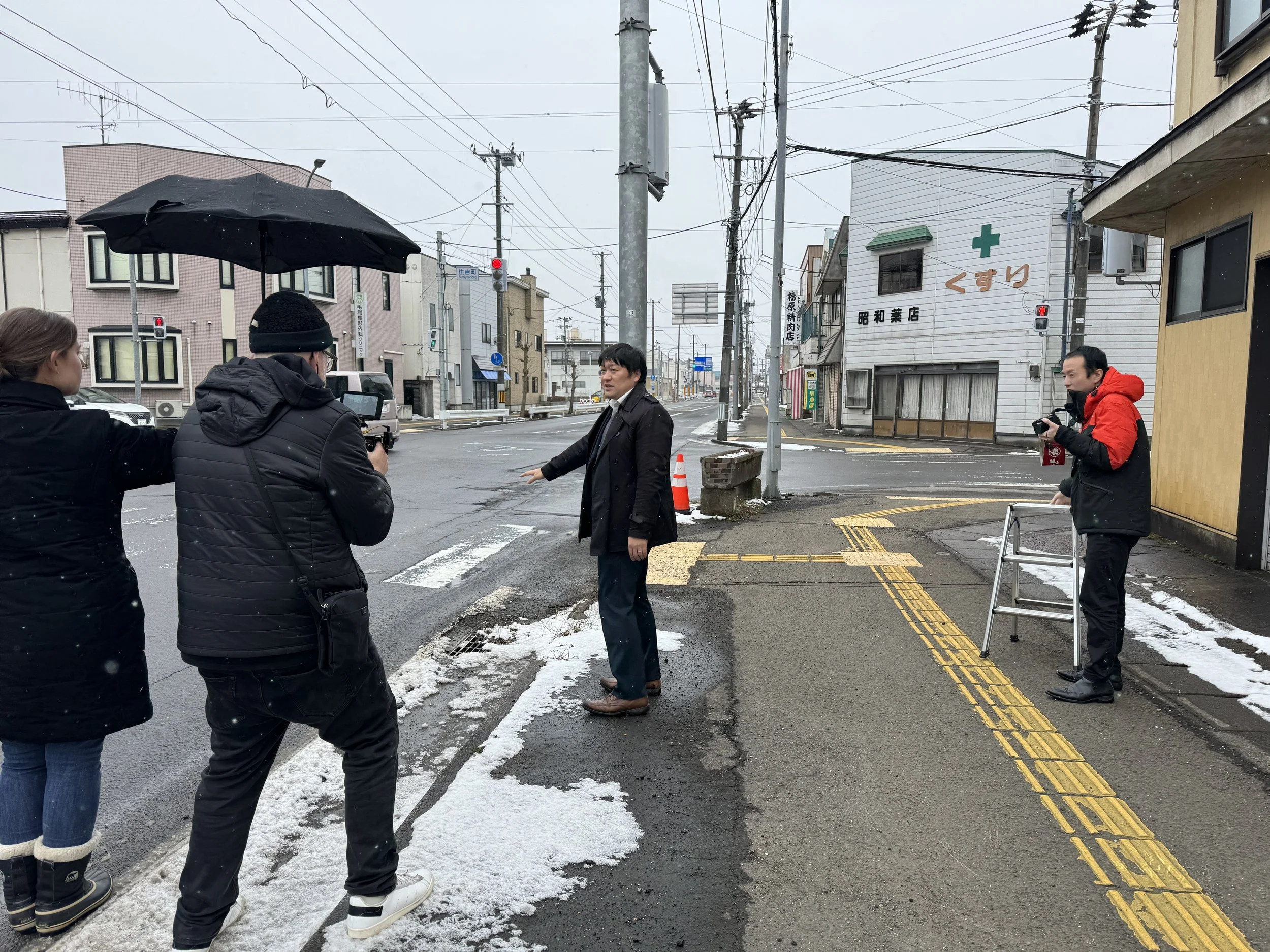 People filming an interview on a snowy city street with electrical wires, buildings, and traffic signals.