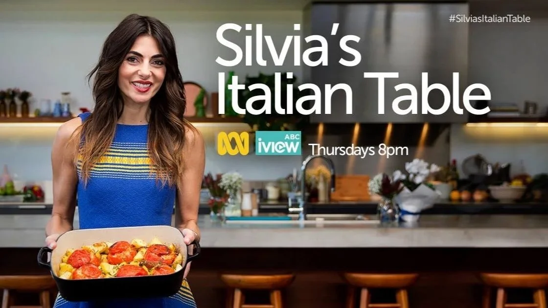 A woman holding a baking dish filled with baked tomatoes and potatoes in a modern kitchen.