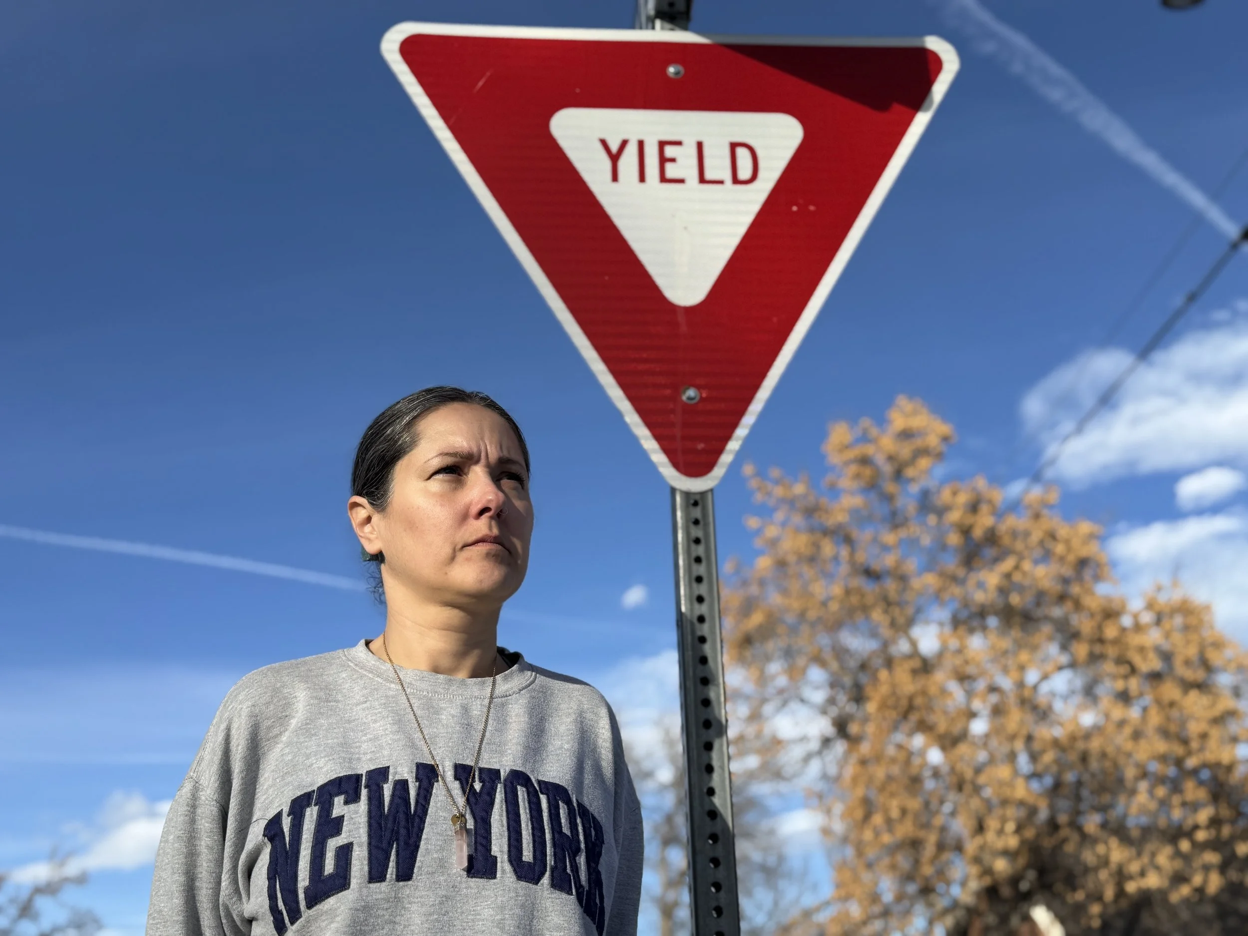 A woman with dark hair pulled back, standing outdoors on a sunny day, looking slightly to the side, in front of a red and white yield traffic sign. She is wearing a gray sweatshirt with the word "New York" on it and a necklace. In the background, blurred trees with autumn-colored leaves can be seen against a blue sky with some clouds.