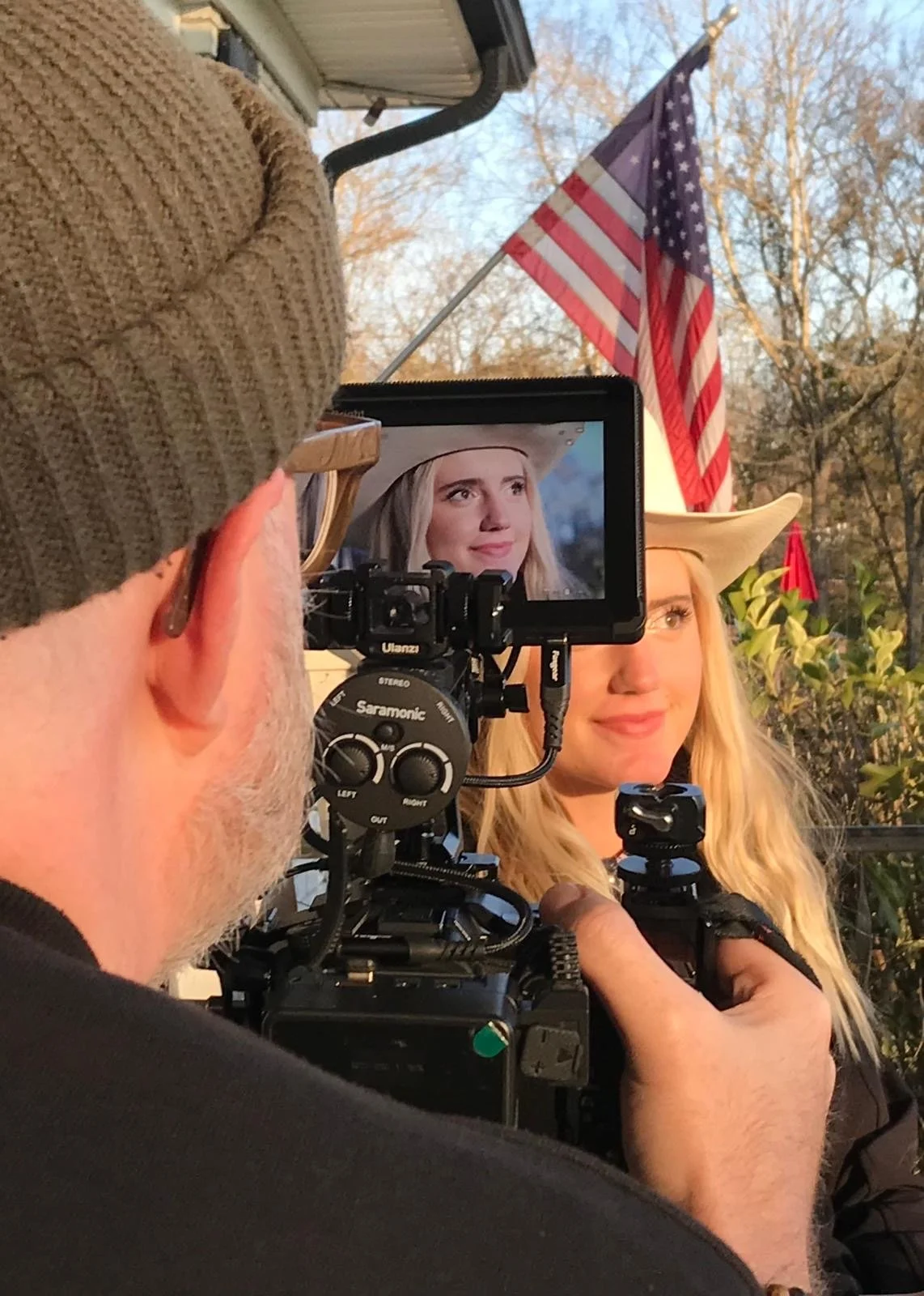 A filmmaker captures a woman wearing a cowboy hat in front of an American flag, outdoors during sunset.