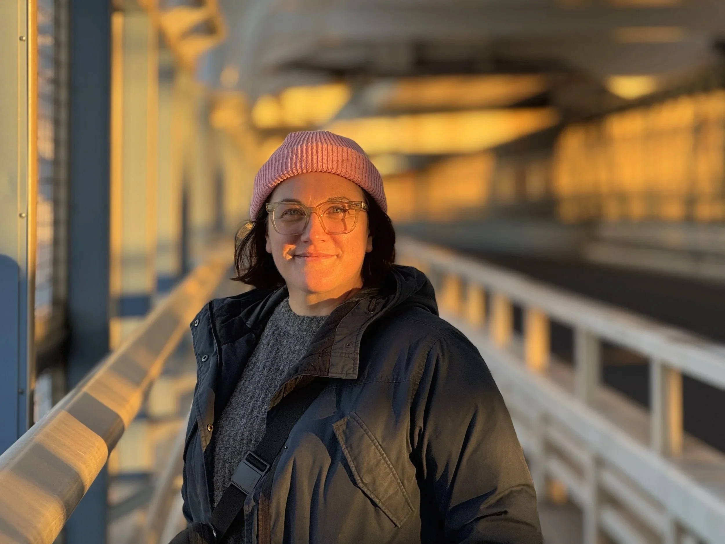 A woman with glasses, wearing a pink beanie and a dark jacket, smiling on a bridge during sunset.