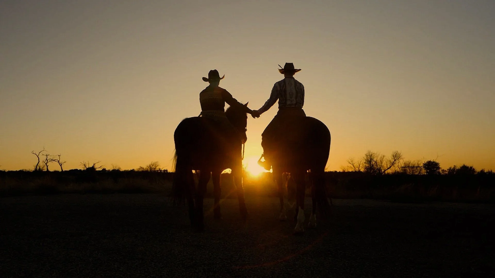 Two cowboys riding horses and holding hands at sunset in a rural landscape with sparse trees.