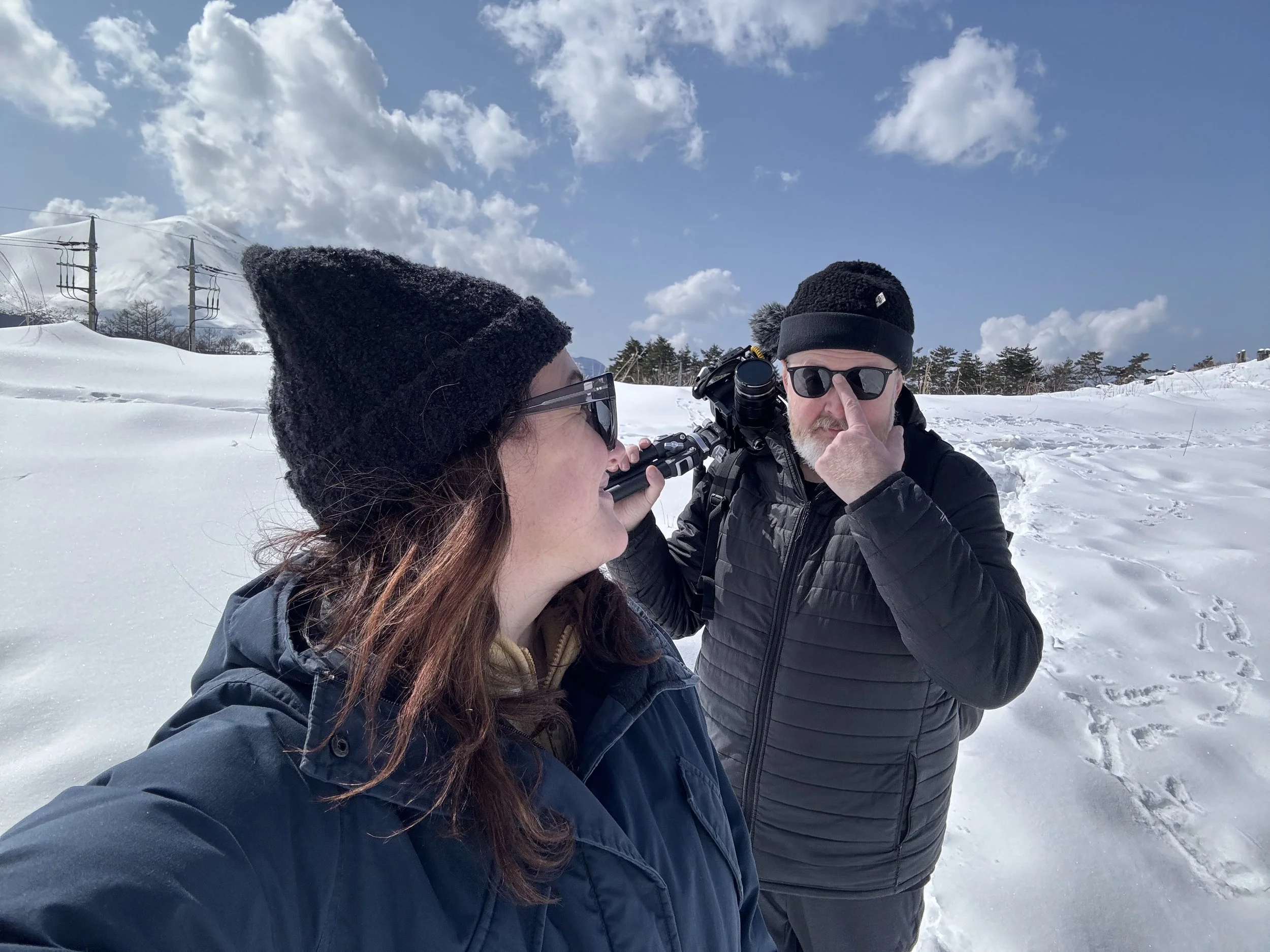 Two people, one woman and one man, in winter clothing and sunglasses in a snowy outdoor setting with cloudy blue sky and electrical poles in the background.
