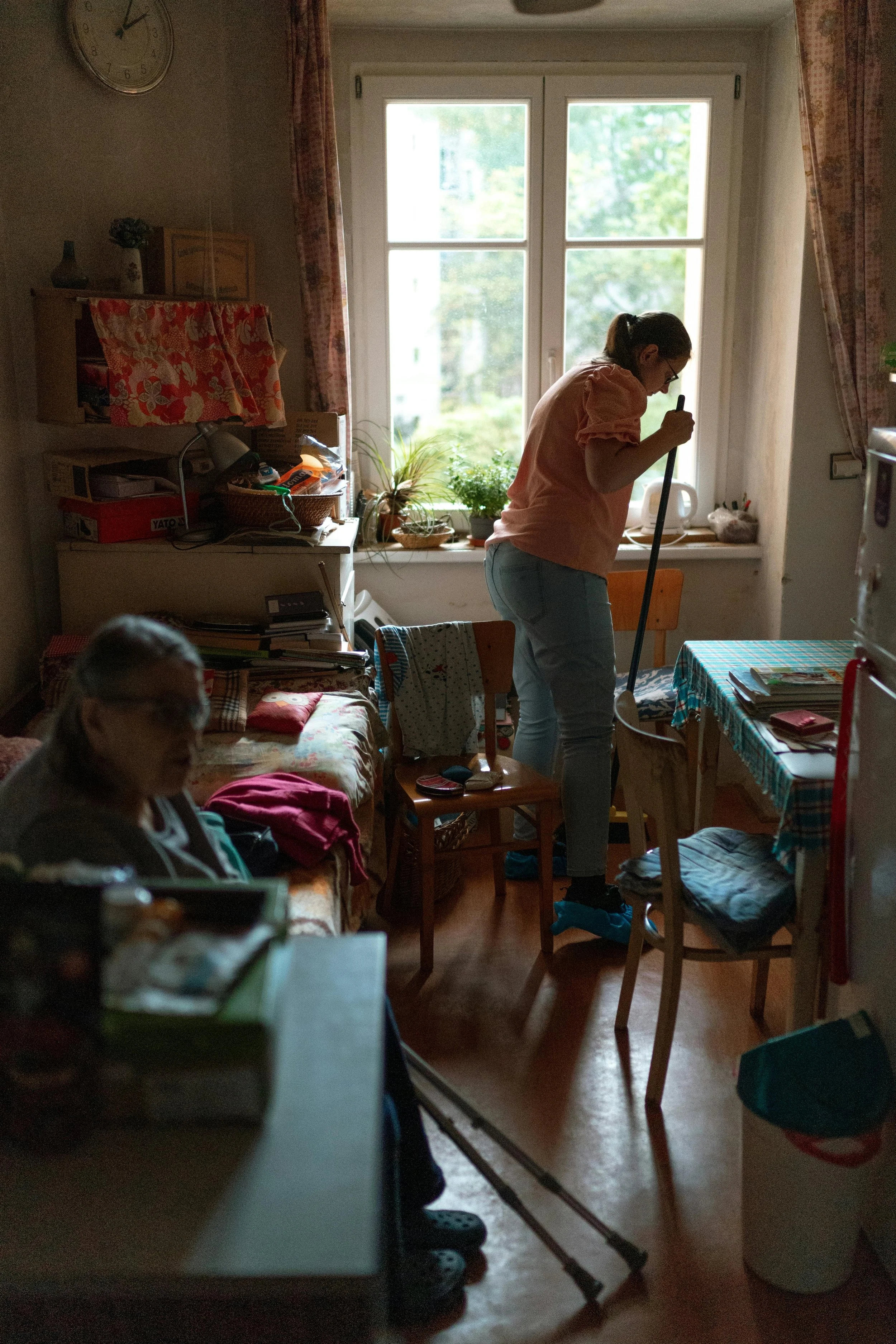 Innenaufnahme eines Zimmers mit einem großen Fenster, jemand arbeitet mit einem Staubwedel, im Vordergrund eine andere Person sitzt auf einem Sofa. Der Raum ist cluttered mit Büchern, Pflanzen und Haushaltsgegenständen.