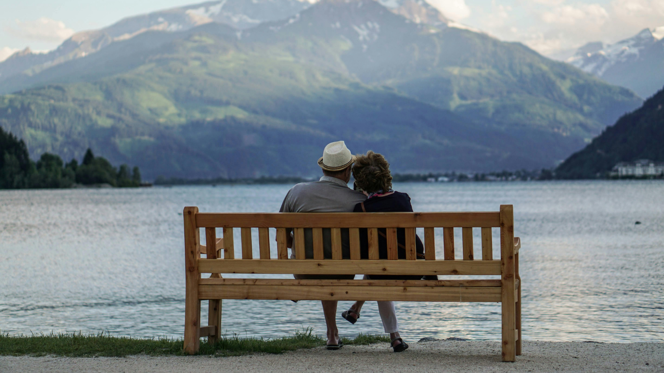 Ein älteres Paar sitzt auf einer Holzbank am Ufer eines Sees mit Blick auf Berge im Hintergrund, vermutlich beim Spaziergang in der Natur.