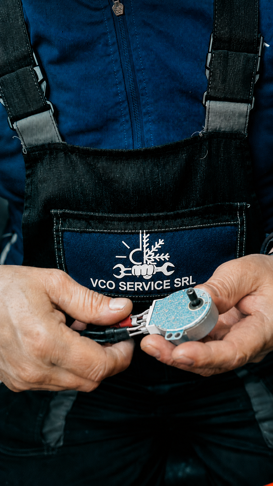 L'immagine mostra un uomo che tiene un motore elettrico di piccole dimensioni con cavi, vestito in tuta da lavoro blu, con una pettorina nera con etichetta "VCO SERVICE SRL".