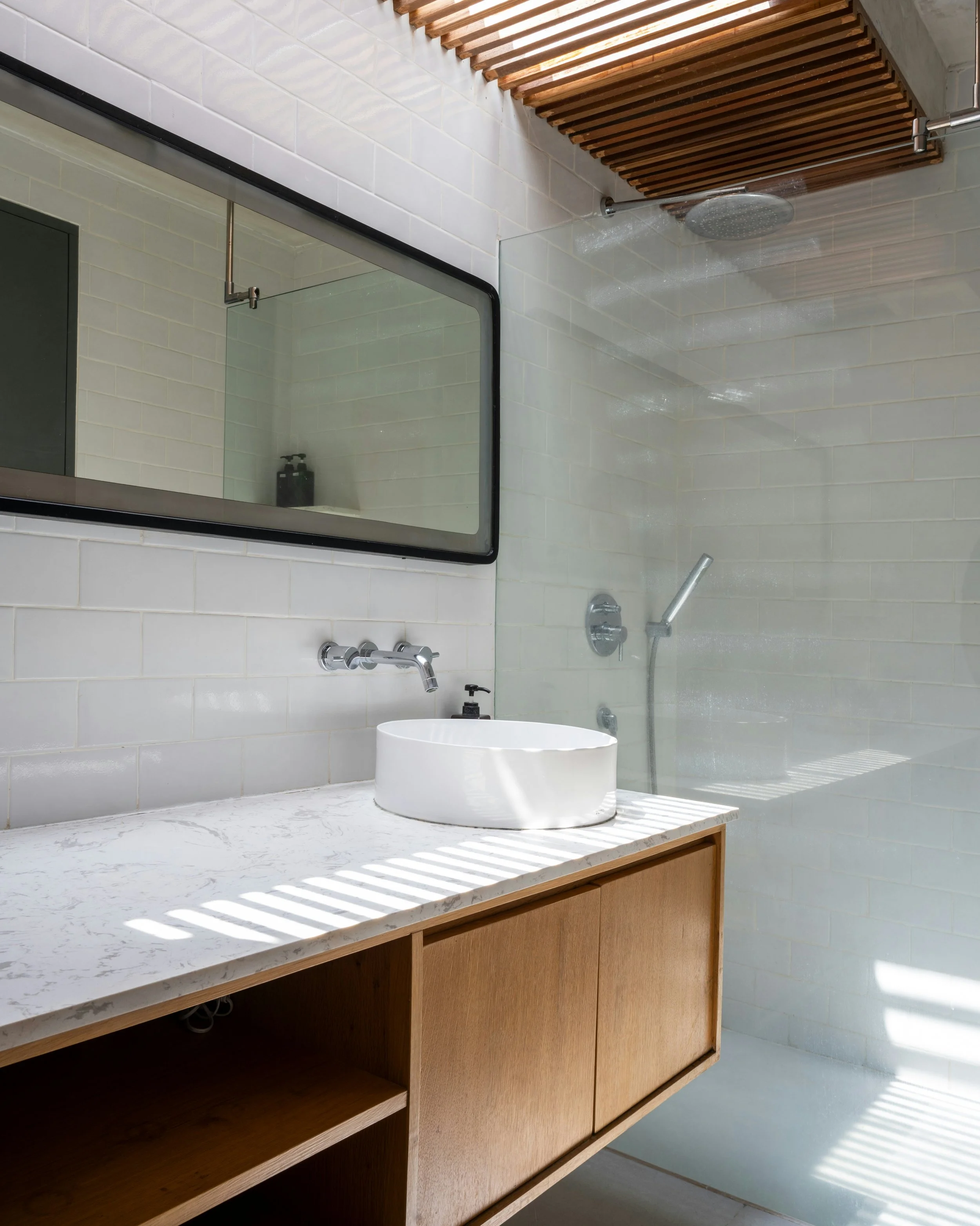 Bagno moderno con lavabo rotondo bianco su un mobile in legno chiaro, specchio grande sulla parete, doccia con soffione a pioggia e pareti in piastrelle bianche, con luci che creano ombre sul piano del lavabo.