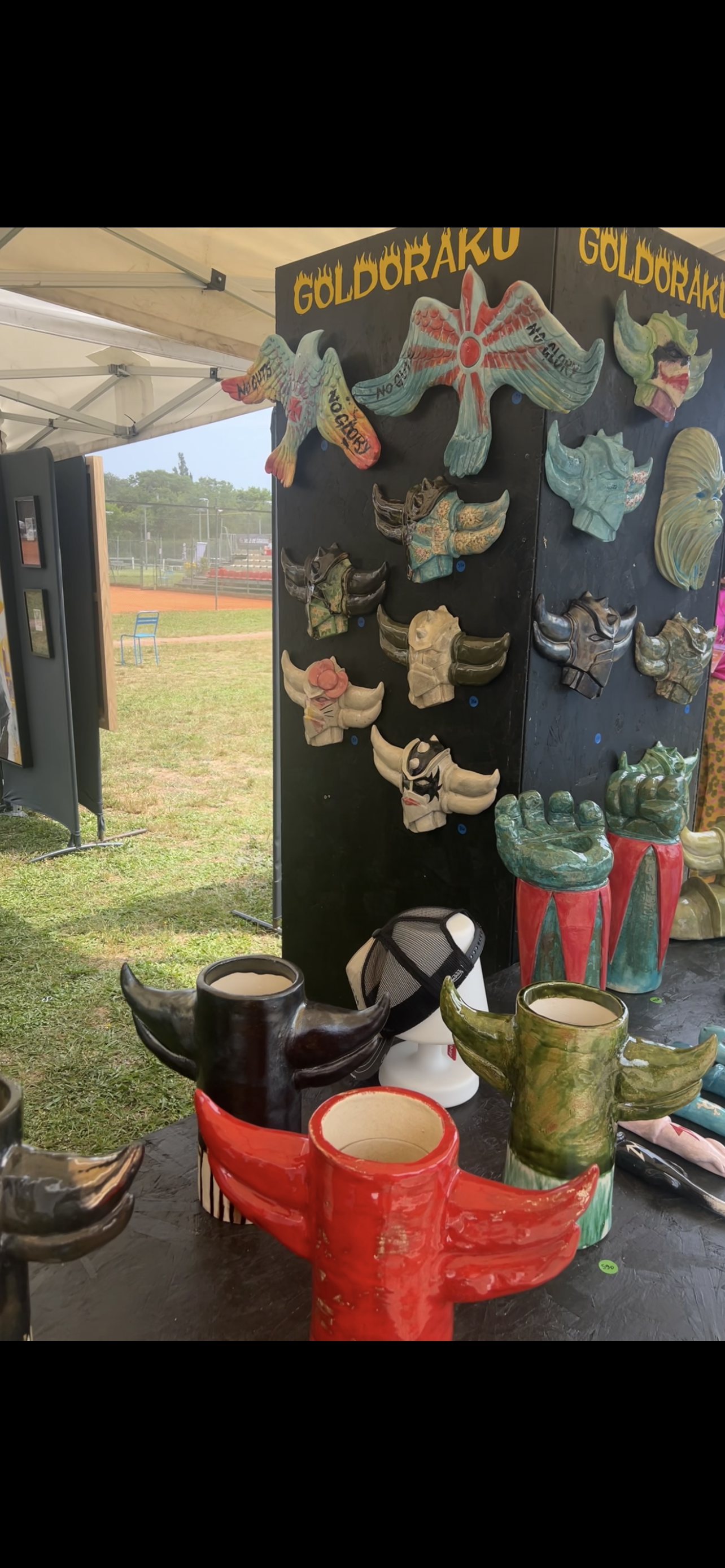 Exposition de masques en bois peint à l'extérieur, avec des masques aux formes de têtes d'animaux colorés et stylisés, et des mugs en forme de têtes d'animaux, notamment des bulles avec des cornes.
