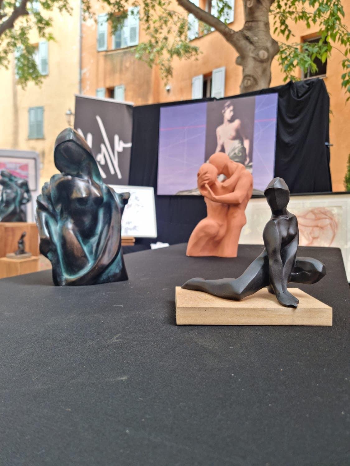 sculptures en exhibition, incluant une tête abstraite en bronze, une sculpture en terre cuite représentant un couple embrassé, et une femme allongée en bronze sur une base en bois, avec un fond montrant une photographie d'un homme nu, dans un cadre d'exposition extérieure.