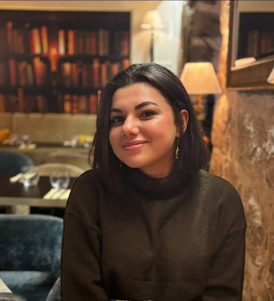 Une femme aux cheveux noirs, portant un pull noir, assise dans un restaurant ou une bibliothèque, avec des murs en pierre, des lampes et des étagères remplies de livres en arrière-plan.