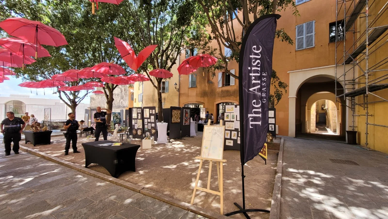 Une cour extérieure avec des arbres et des parasols rouges suspendus. Il y a une exposition d'art avec des panneaux et des œuvres, des tables avec des documents, et une bannière noire avec l'inscription "The Artiste GRASSE". Un passage sous un bâtiment orange avec des fenêtres à volets bleus et une échelle de scaffolding à droite.