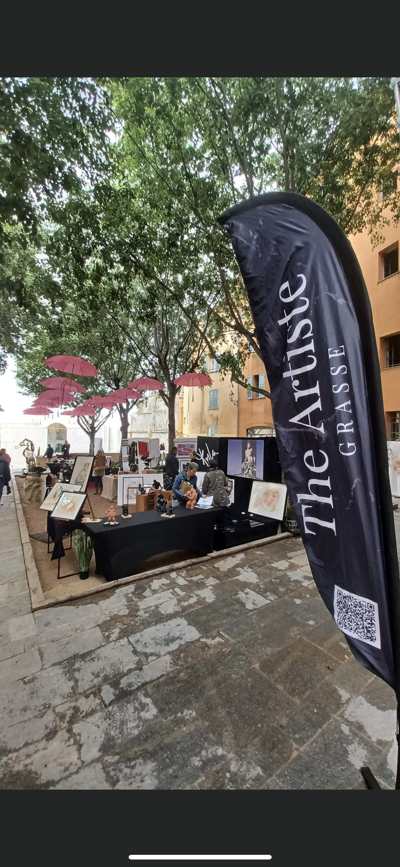 Marché d'artisanat en plein air avec stands d'objets d'art et d'artisanat, parasols roses suspendus dans un parc entouré d'arbres et de bâtiments antigais