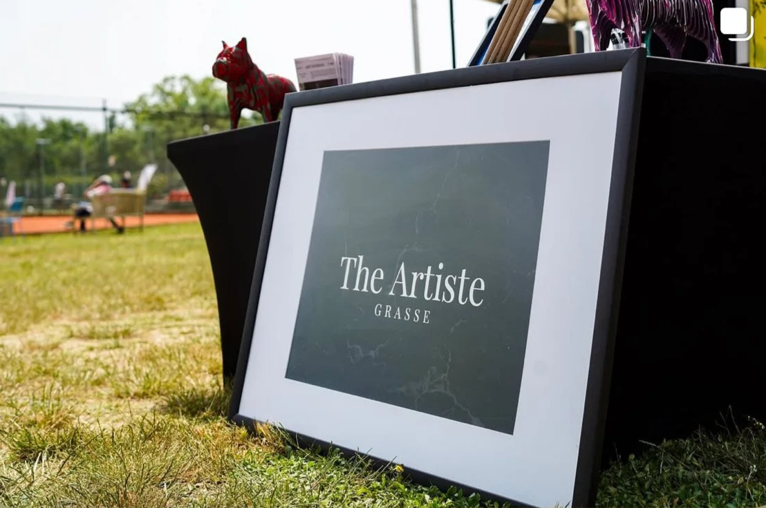 Tableau avec le texte : The Artiste GRASE, positionné sur une surface extérieure avec un sol en herbe, en face d'un terrain de tennis ou de sport.