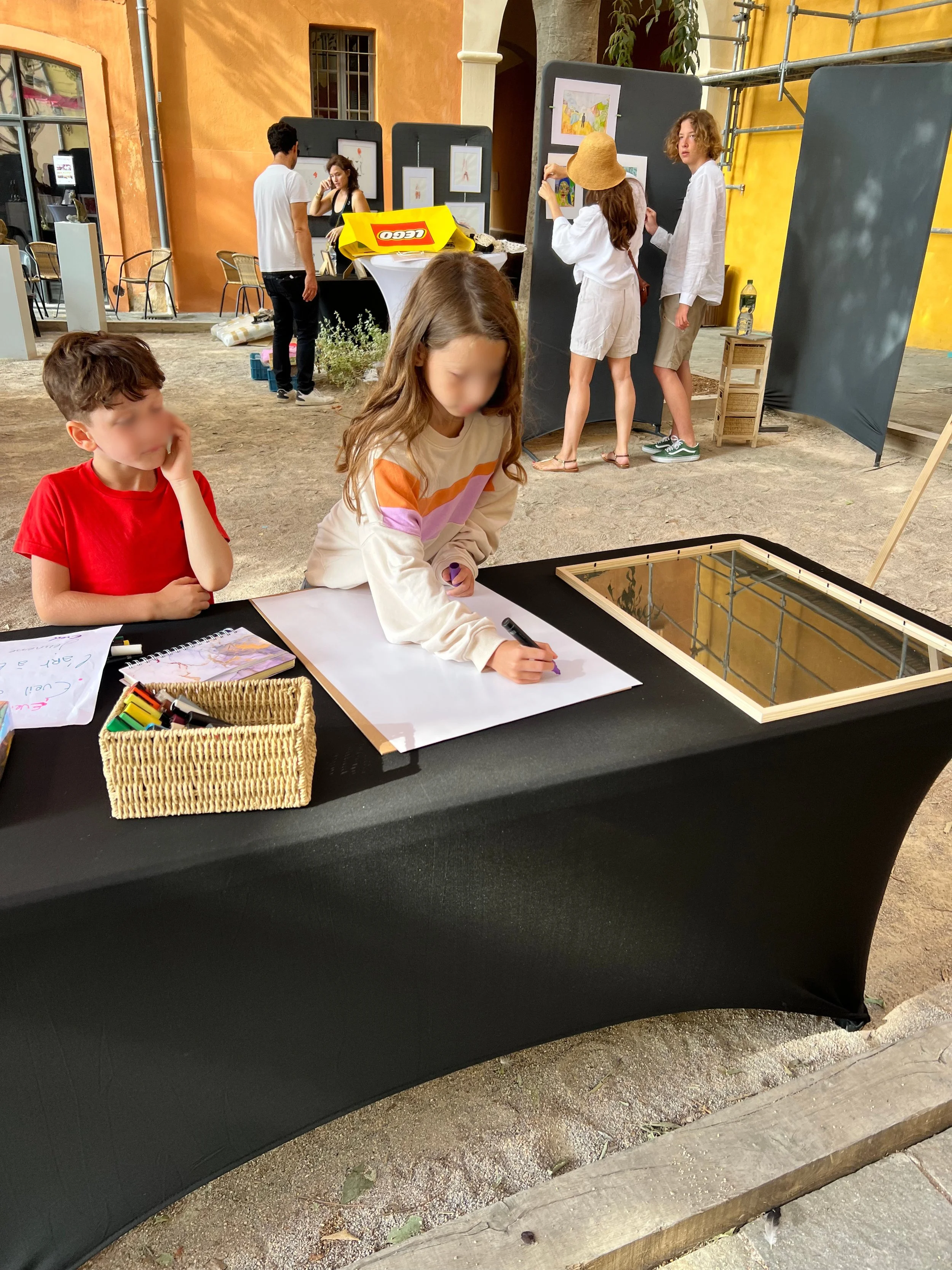 Une scène de rue ou d'événement artistique avec enfants et adultes. Deux enfants sont assis à une table avec des crayons et du papier, un garçon en rouge et une fille en blanc avec des bandes orange et violette. En arrière-plan, des adultes regardent des œuvres d'art affichées sur des panneaux, et un groupe de personnes discute ou observe. Le sol est en terre battue, et il y a des structures métalliques et un bâtiment en jaune-orange en arrière-plan.