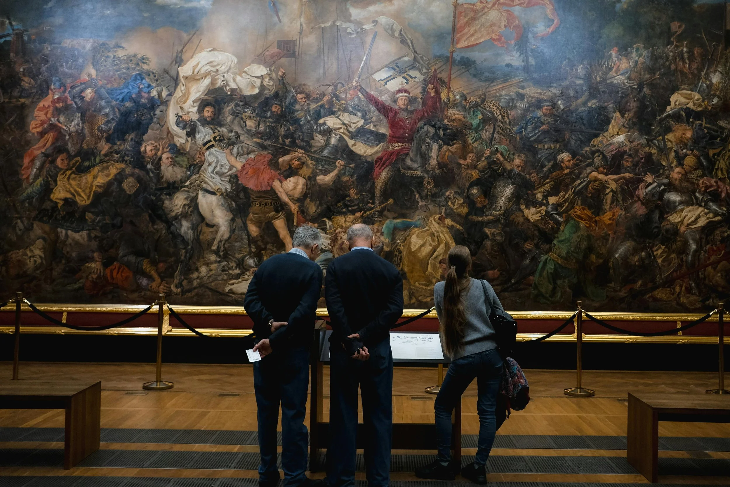 Peinture historique représentant une bataille, exposée dans un musée, avec plusieurs visiteurs regardant l'œuvre.