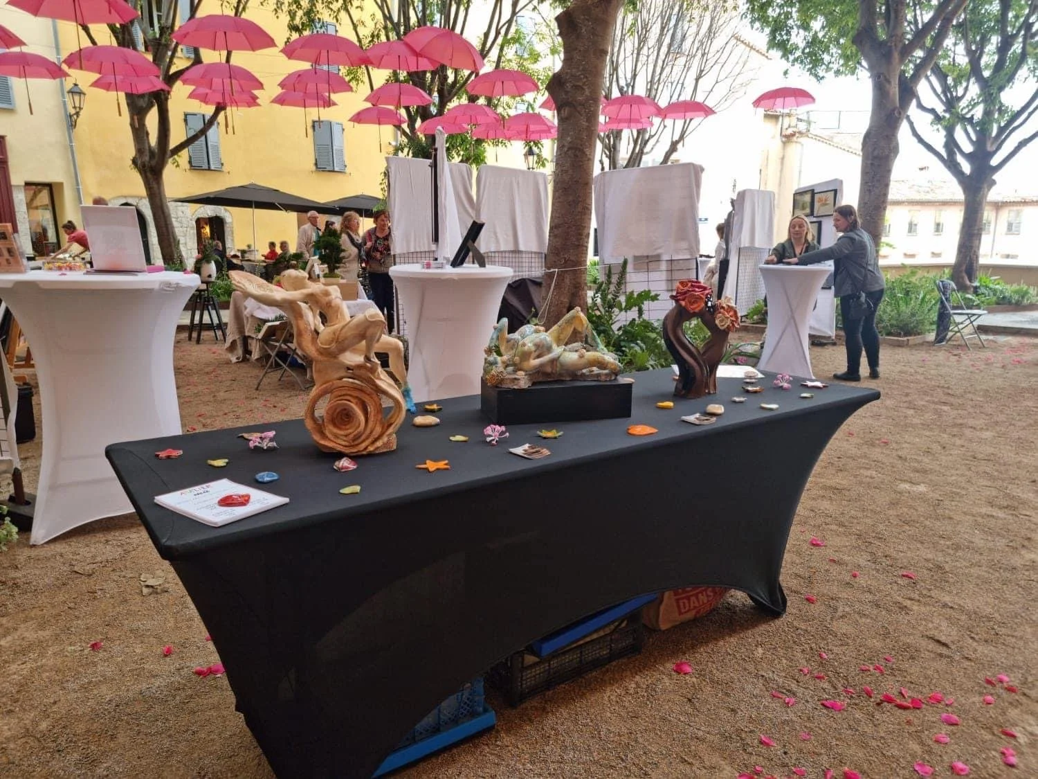Une scène de marché ou d'exposition en plein air avec plusieurs tables, décorations en sculptures, et des parapluies roses suspendus au-dessus. Des personnes discutent et examinent les objets, dans un environnement en plein air avec des arbres et un bâtiment en arrière-plan.