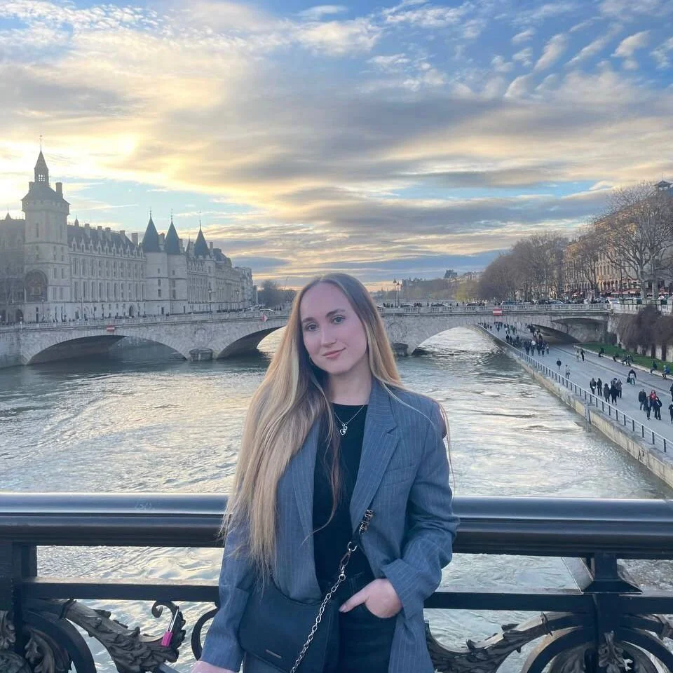 Une jeune femme blonde en veste grise avec une chaîne en or pose devant la Seine à Paris, avec le Château de la Conciergerie et le pont en arrière-plan, lors d'une journée nuageuse.