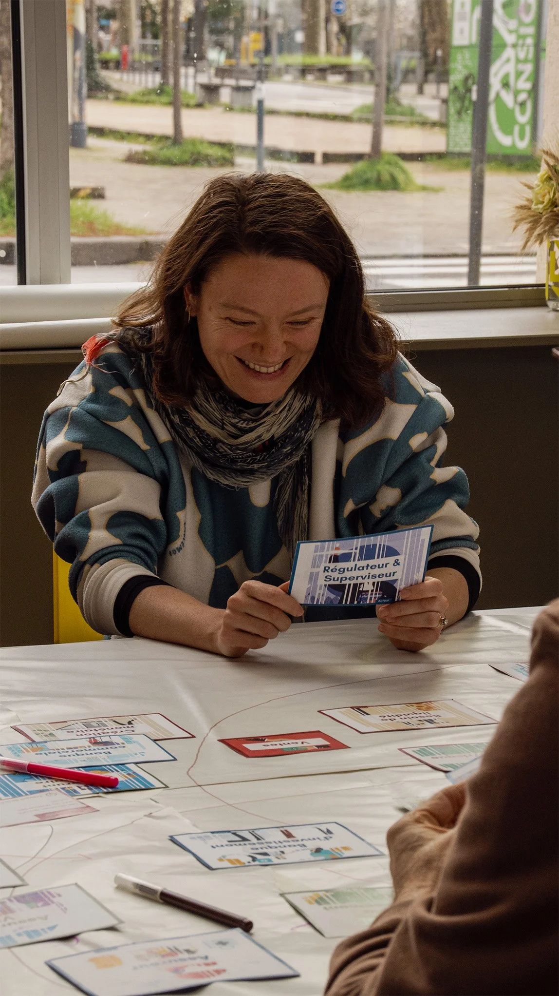 Une femme souriante tient un carte avec l'inscription 'Régulateur & Superviseur' lors d'une réunion ou activité de groupe, autour d'une table avec diverses brochures et un stylo rouge.