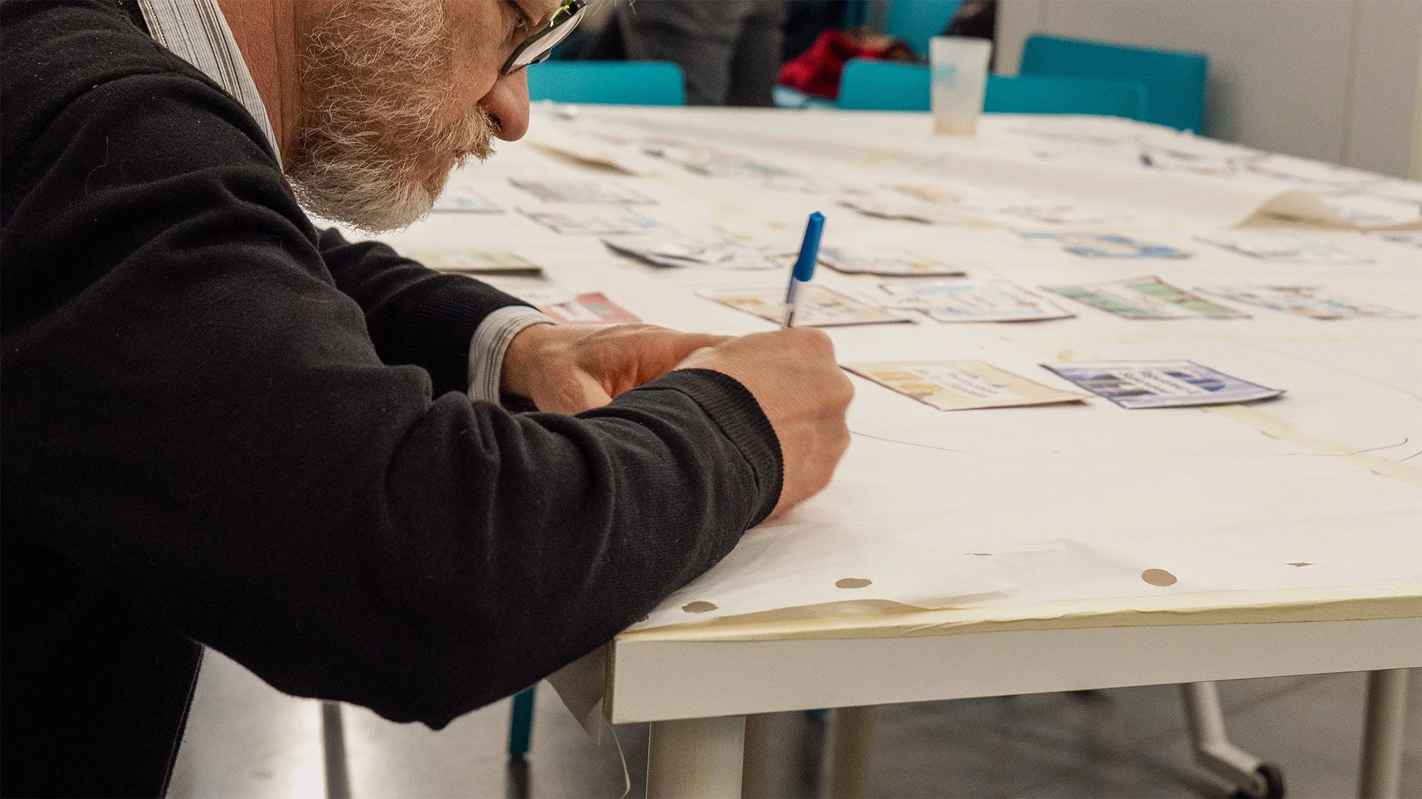 Un homme âgé, portant des lunettes et une barbe blanche, écrit ou dessine dans le cadre d'une fresque.