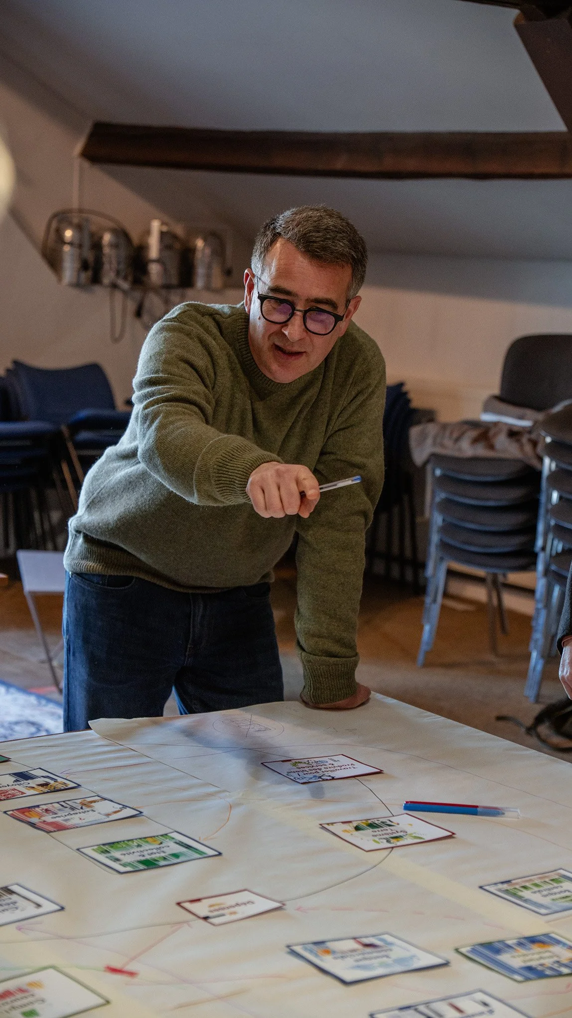 Un homme porte des lunettes, portant un pull vert, qui pointe un objet sur une grande table couverte de cartes de la fresque de la finance.
