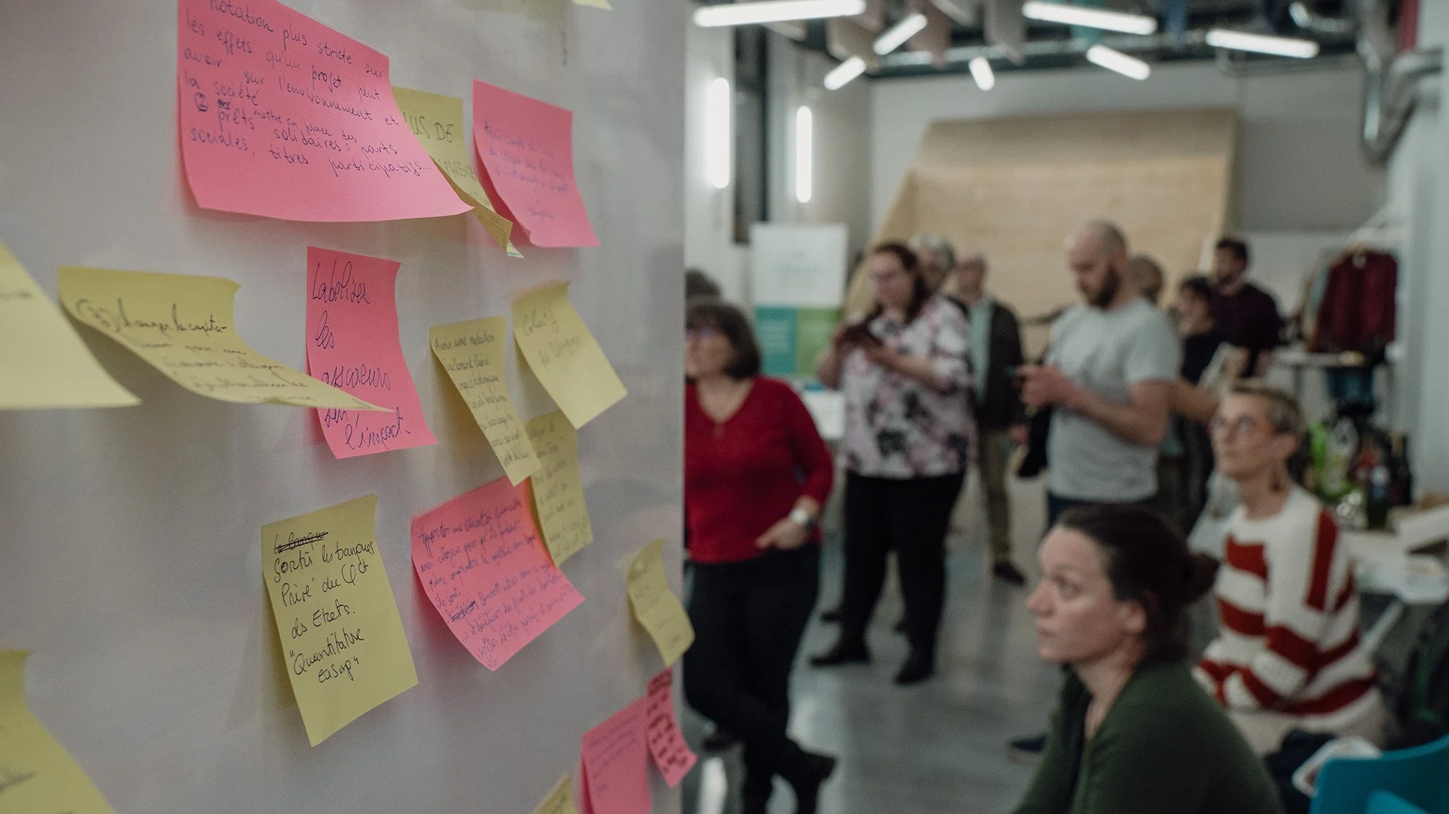 Post-it de différentes couleurs avec des écrits en français sur un mur, un groupe de personnes discutant pendant un atelier.