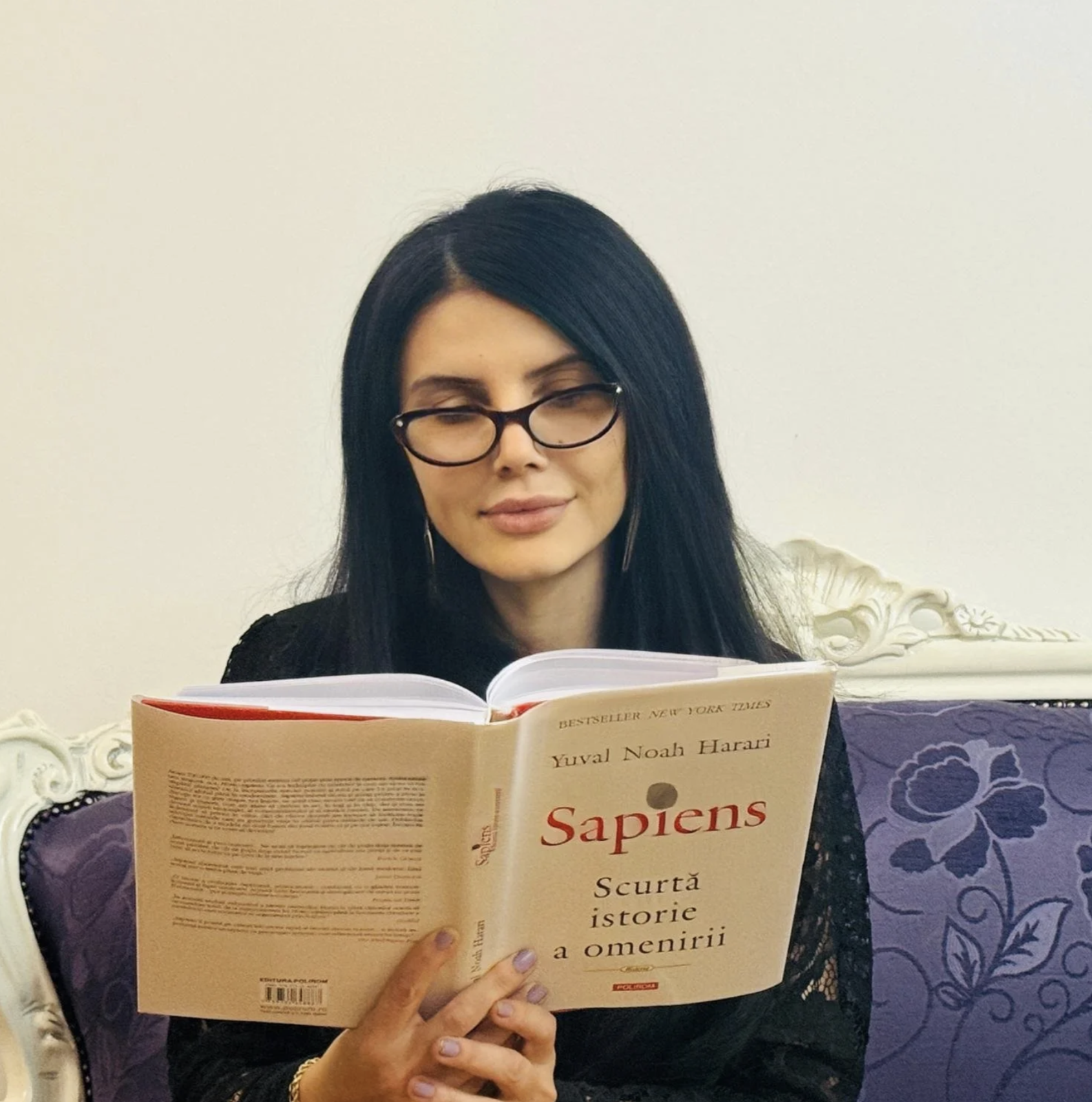 A woman with black hair, glasses, and light makeup reading a book titled 'Sapiens' by Yuval Noah Harari while sitting on a purple ornate chair.