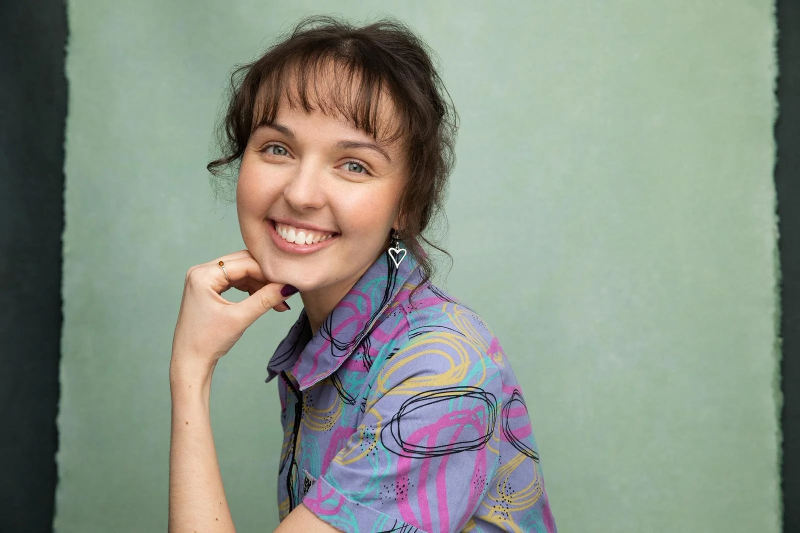 A woman with short, brown hair and light skin smiling, wearing a colorful, patterned shirt and heart-shaped earrings, with her hand resting on her chin, against a green background.