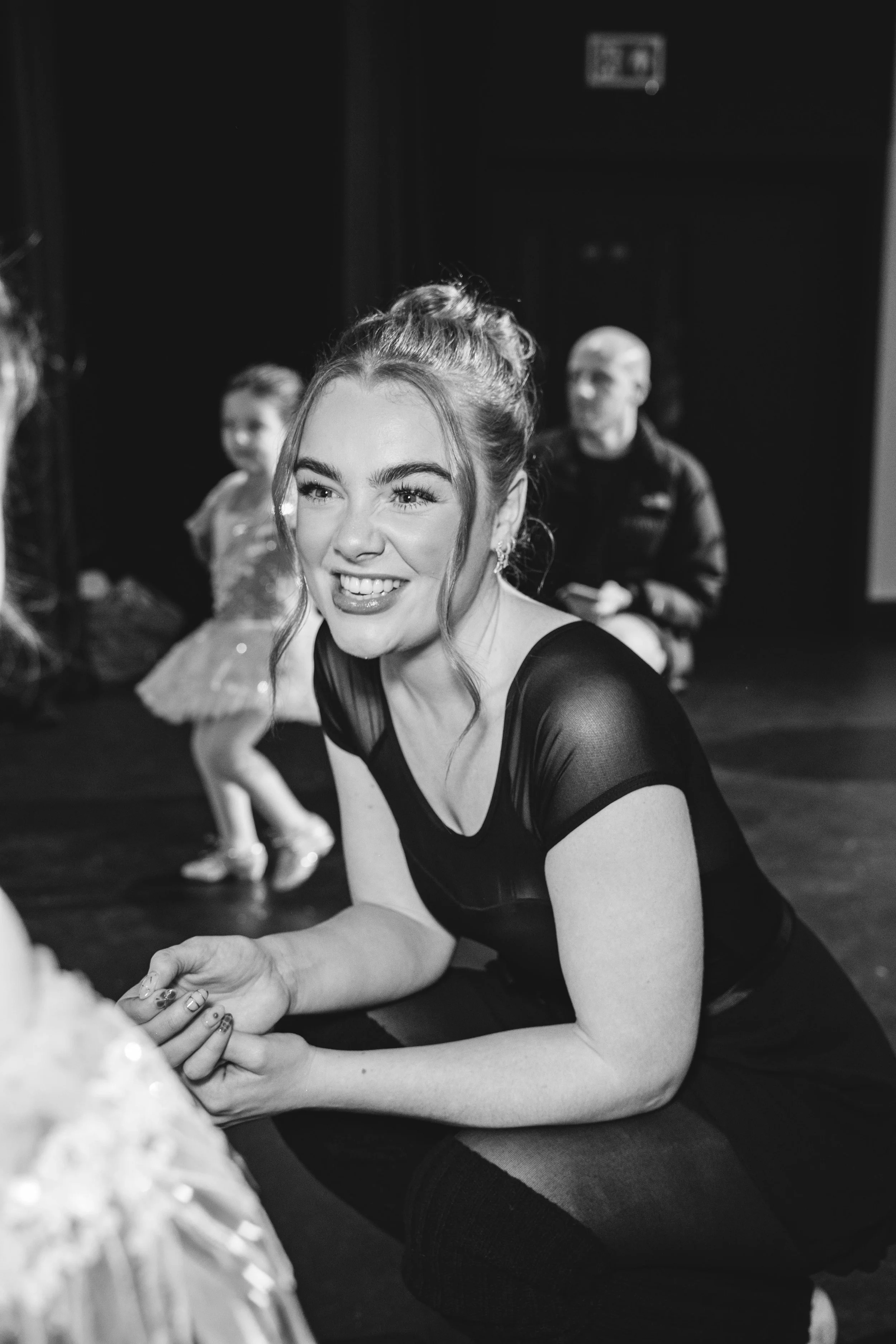 A smiling brunette woman with braided hair, wearing a black dress with sheer sleeves, is sitting on the floor in a dance studio, leaning on her hands, in a dance pose.