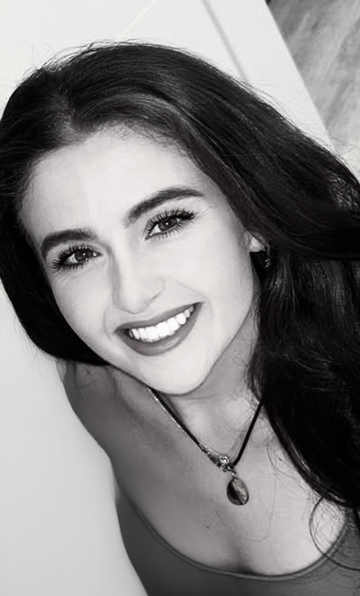 A portrait of a smiling woman with dark hair and eyes, wearing a necklace with a pendant.