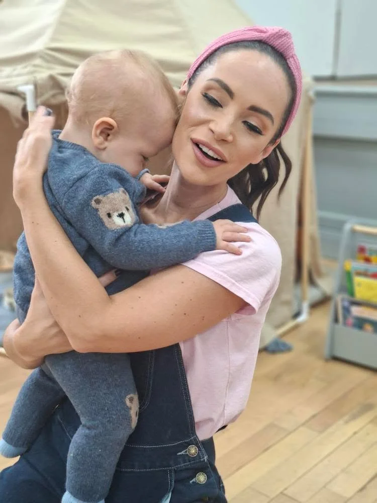 A woman with a pink headband holding and hugging a young child wearing a blue sweater with bear designs, in a room with wooden floors and a bookshelf in the background.