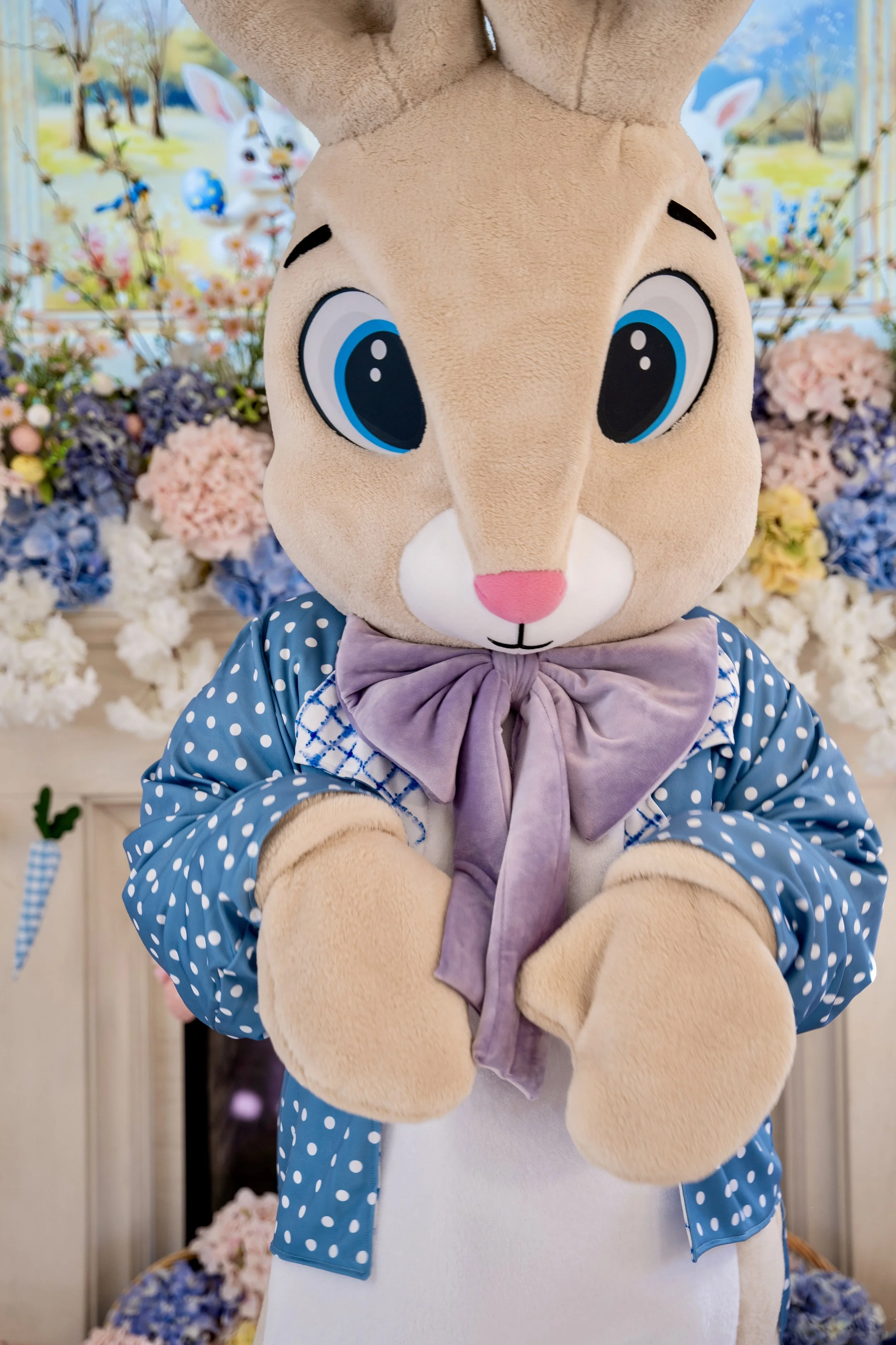A person dressed as a large bunny costume with big blue eyes, pink nose, and beige fur, wearing a blue polka-dot outfit with a lavender bow tie, standing in front of a colorful spring-themed background with flowers and bunny decorations.
