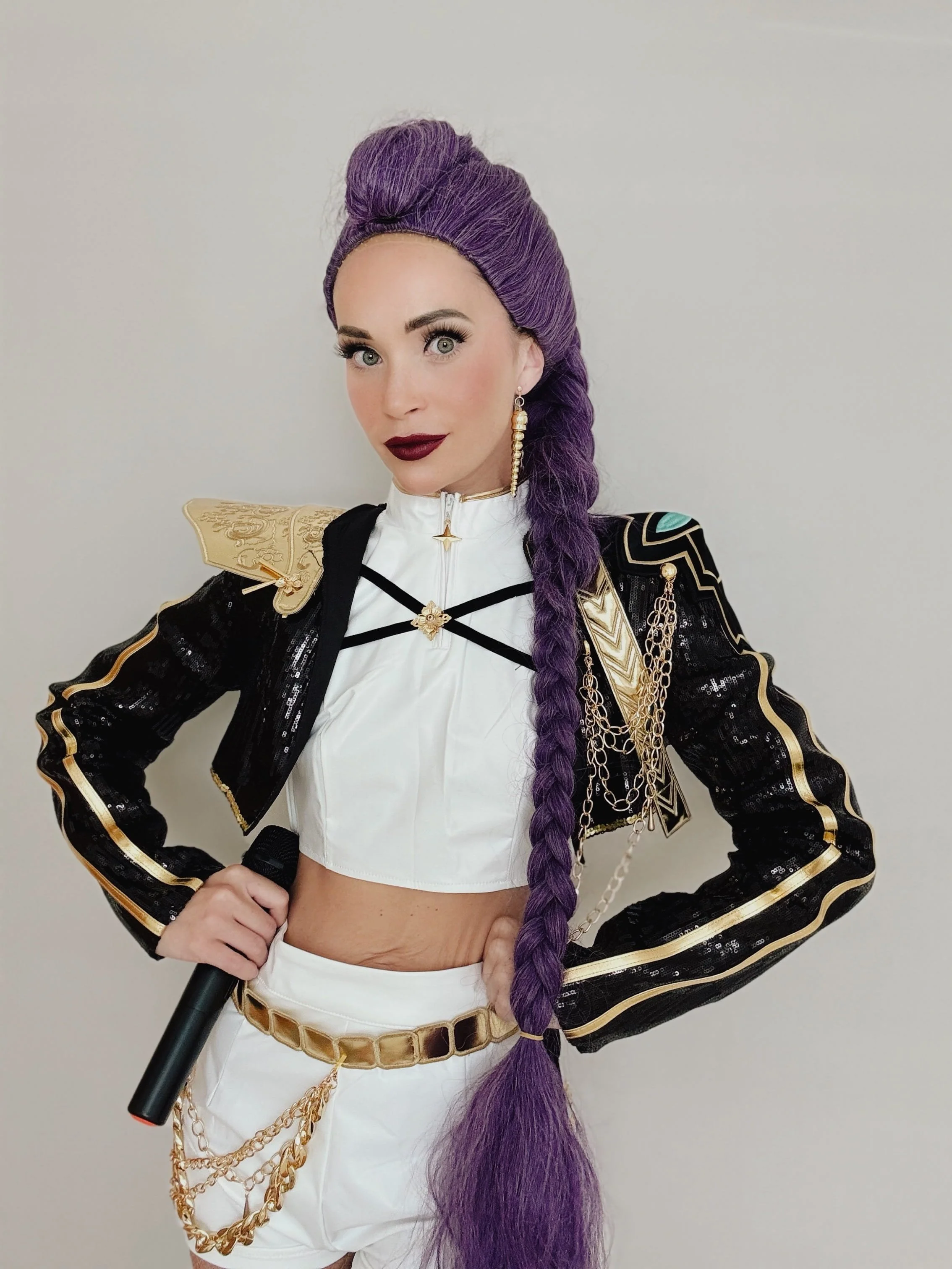 A woman with long purple braided hair, wearing a black and gold military-style jacket and white high-waisted shorts, holding a microphone, standing against a plain gray background.