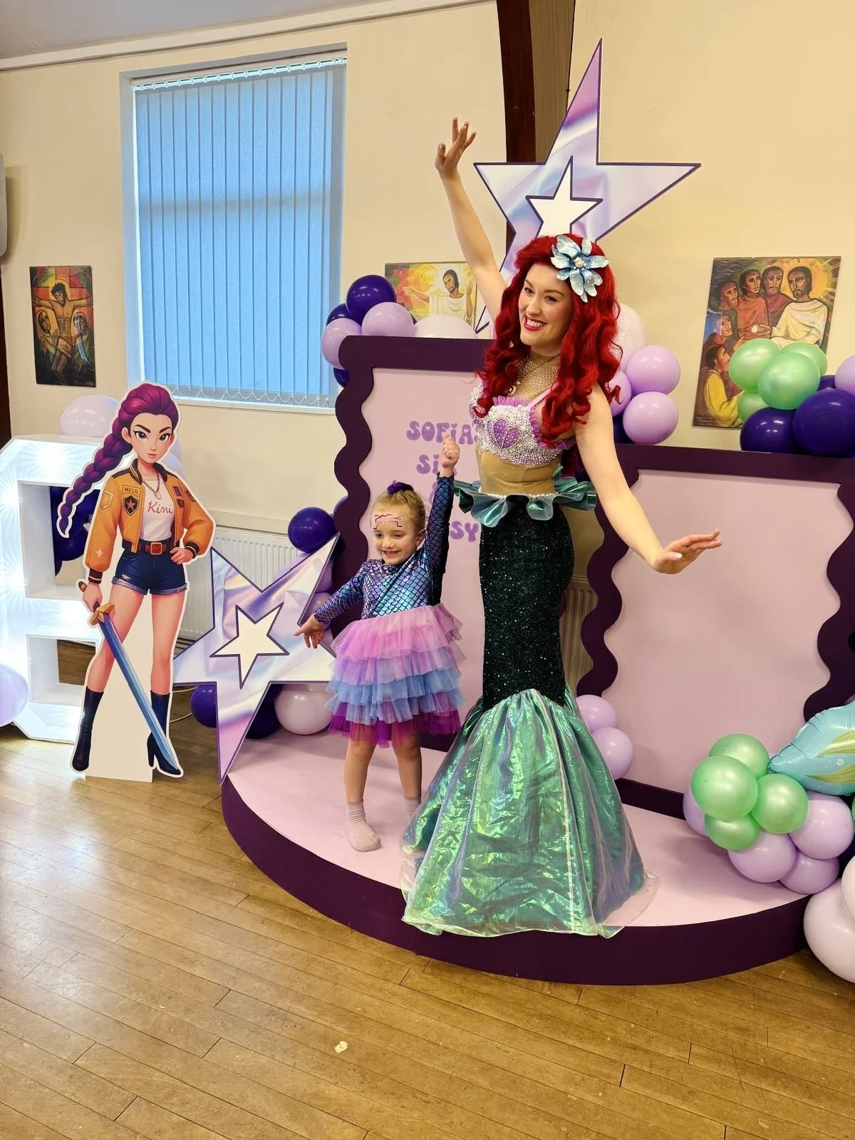 A woman dressed as a mermaid and a young girl in a colorful dress posing at a themed party. The background has balloons, a star decoration, and cartoon character cutouts.